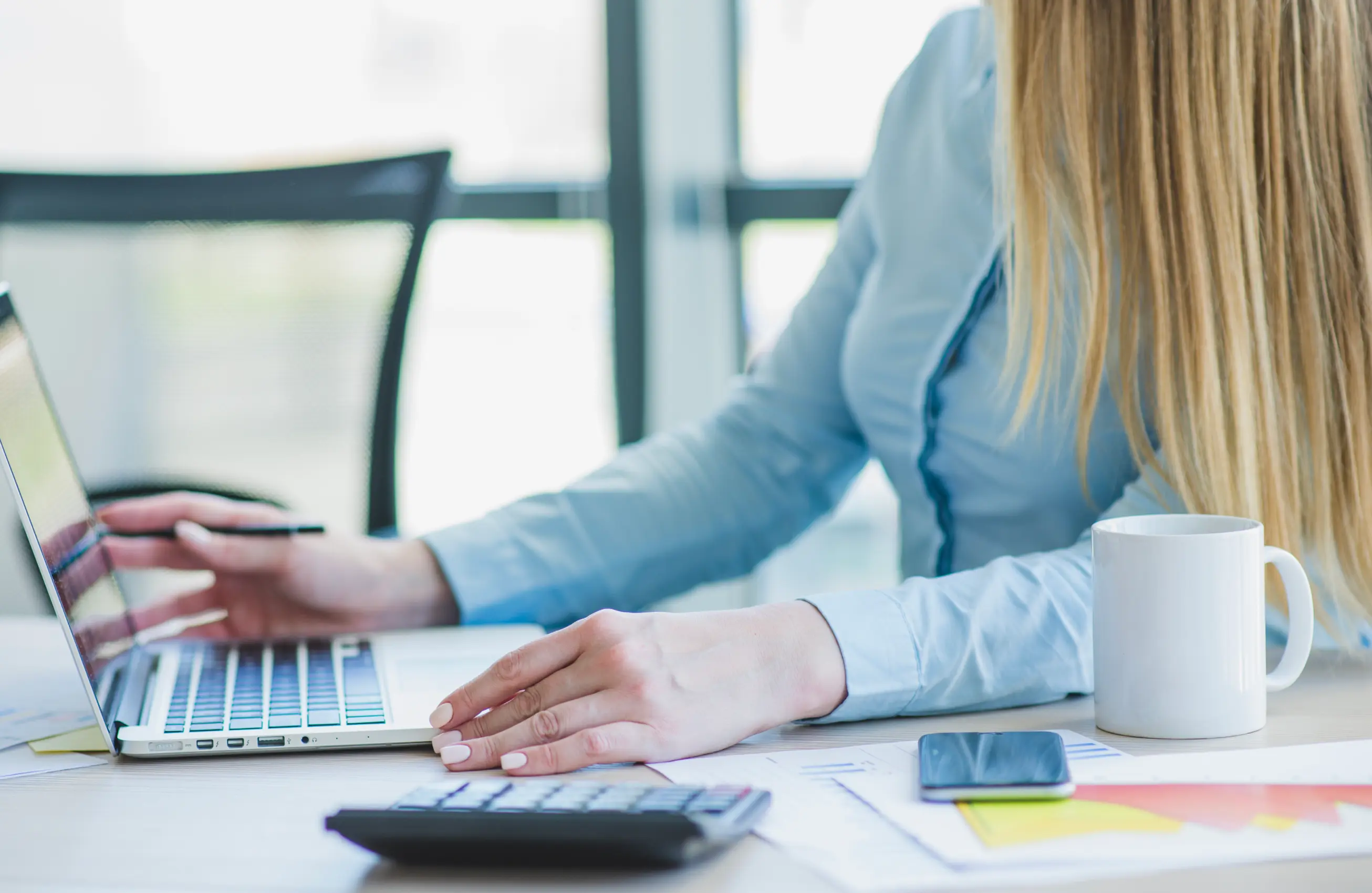 Uma imagem close-up sobre uma mesa com um notebook, onde uma mulher anota algo em uma folha com gráficos enquanto segura um tablet.