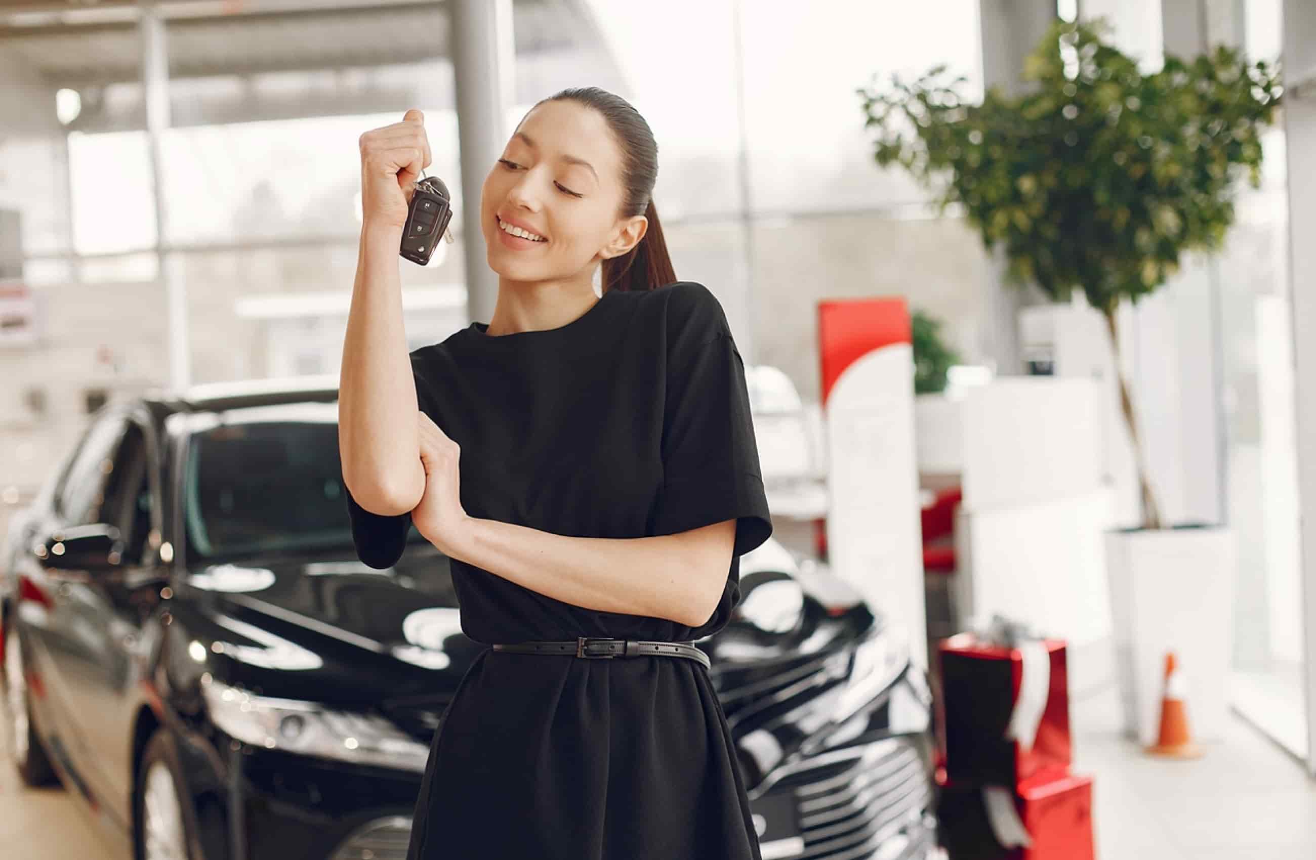Mulher branca com camiseta e calça pretas segurando a chave de um carro. Ao fundo vemos um carro preto dentro de uma concessionária. A imagem representa carros de até 50 mil reais.