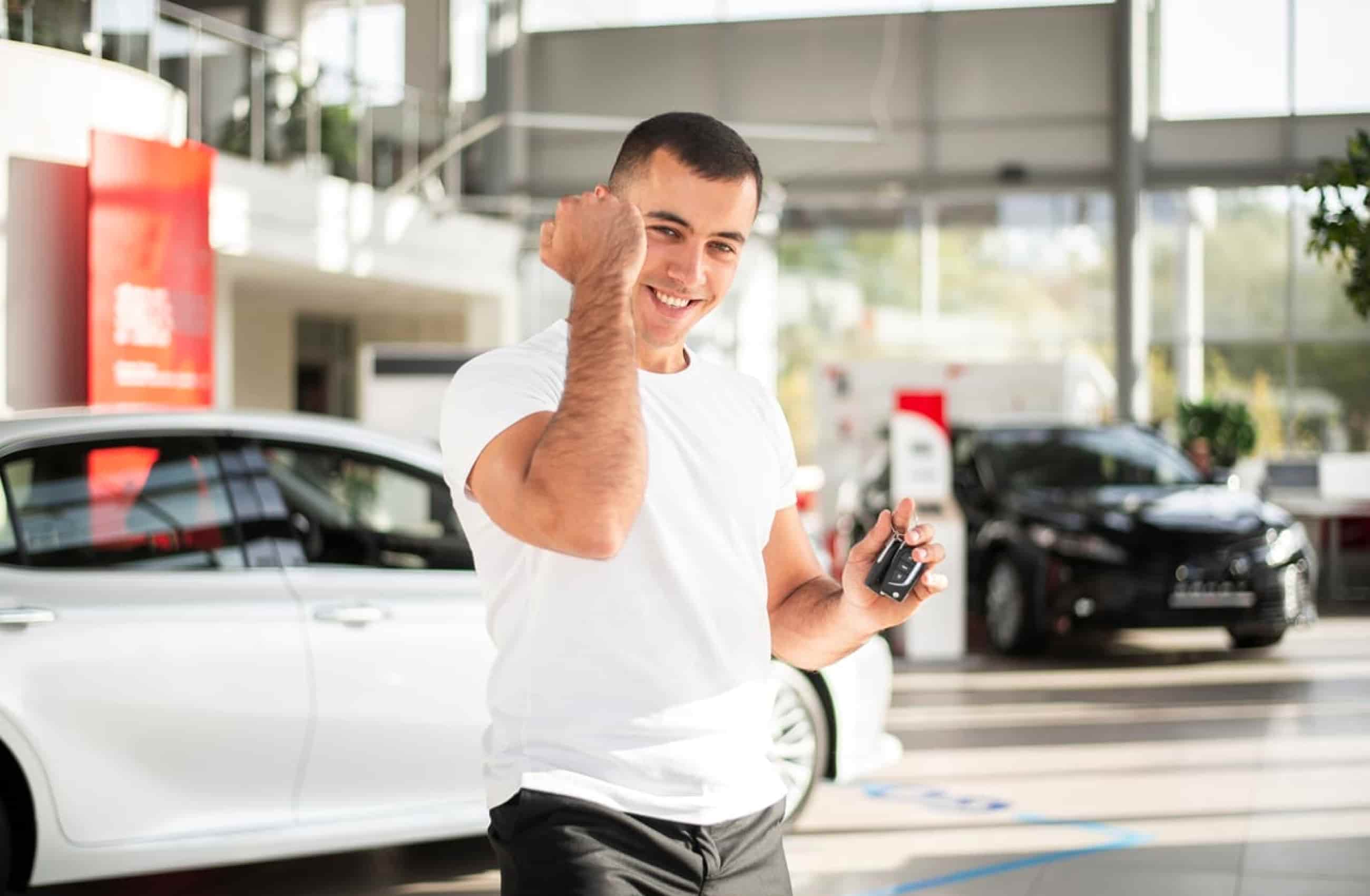 Homem branco com camiseta branca. Ele está segurando uma chave de carro em uma das mãos, e o outro braço faz sinal de comemoração. Ao fundo vemos uma concessionária de carros. A imagem representa veículos alienados.