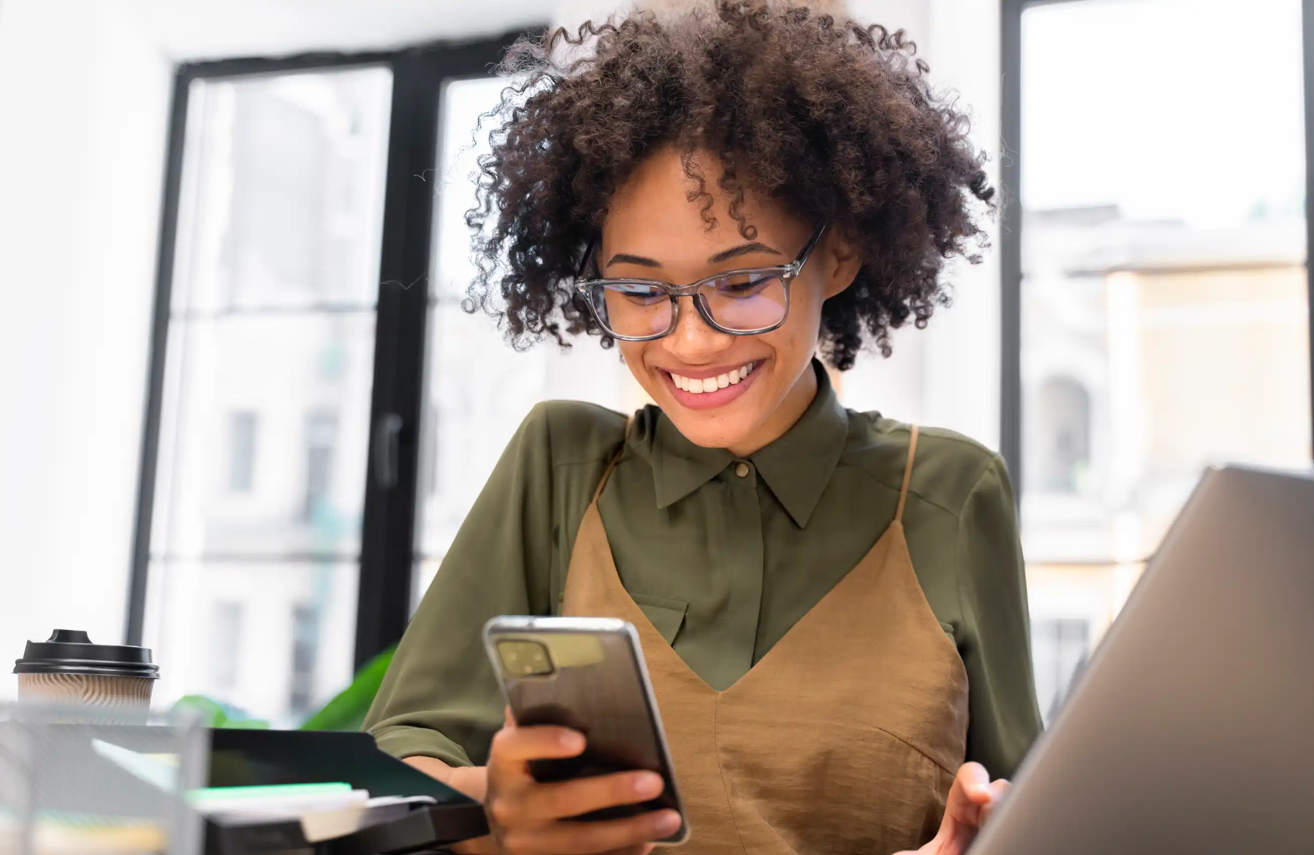 Mulher sorridente sentada à mesa com laptop e copo de café, olhando para o smartphone na mão; ambiente claro de escritório ou coworking.