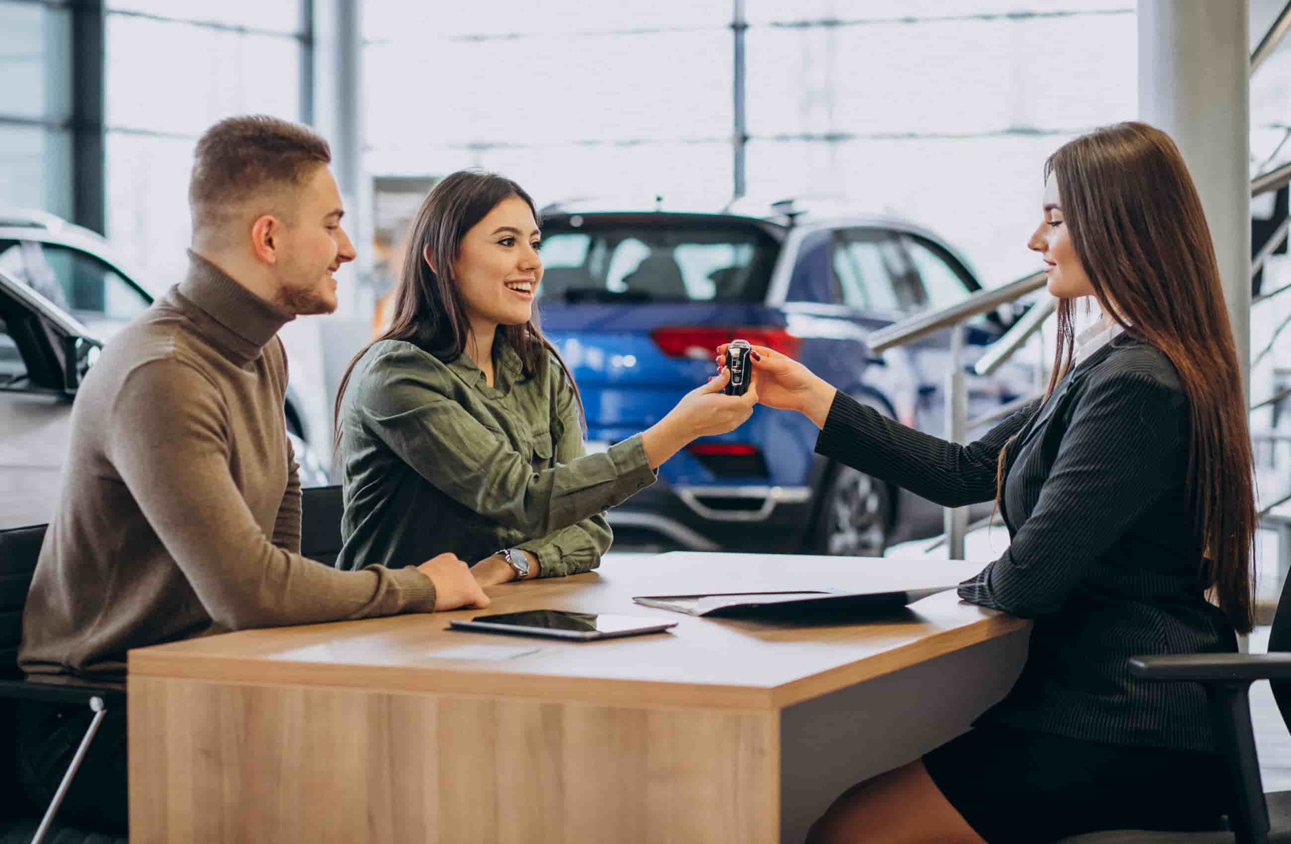 Um homem e uma mulher formam um casal comprando um carro em uma agência de veículos. Ambos estão sentados em frente a uma mesa de madeira, e em sua frente está uma mulher de terno que vende carros segurando uma chave de carro.