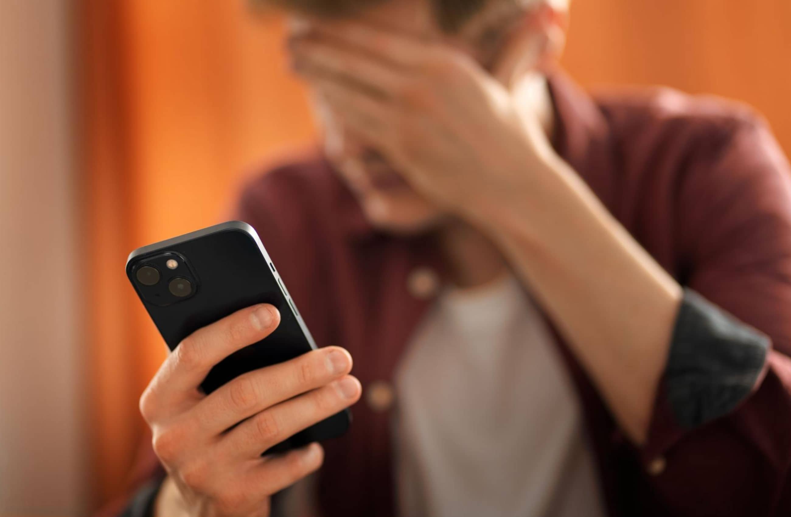 Homem jovem cobrindo o rosto em sinal de estresse enquanto segura um celular, ilustrando o impacto emocional de um golpe do WhatsApp.