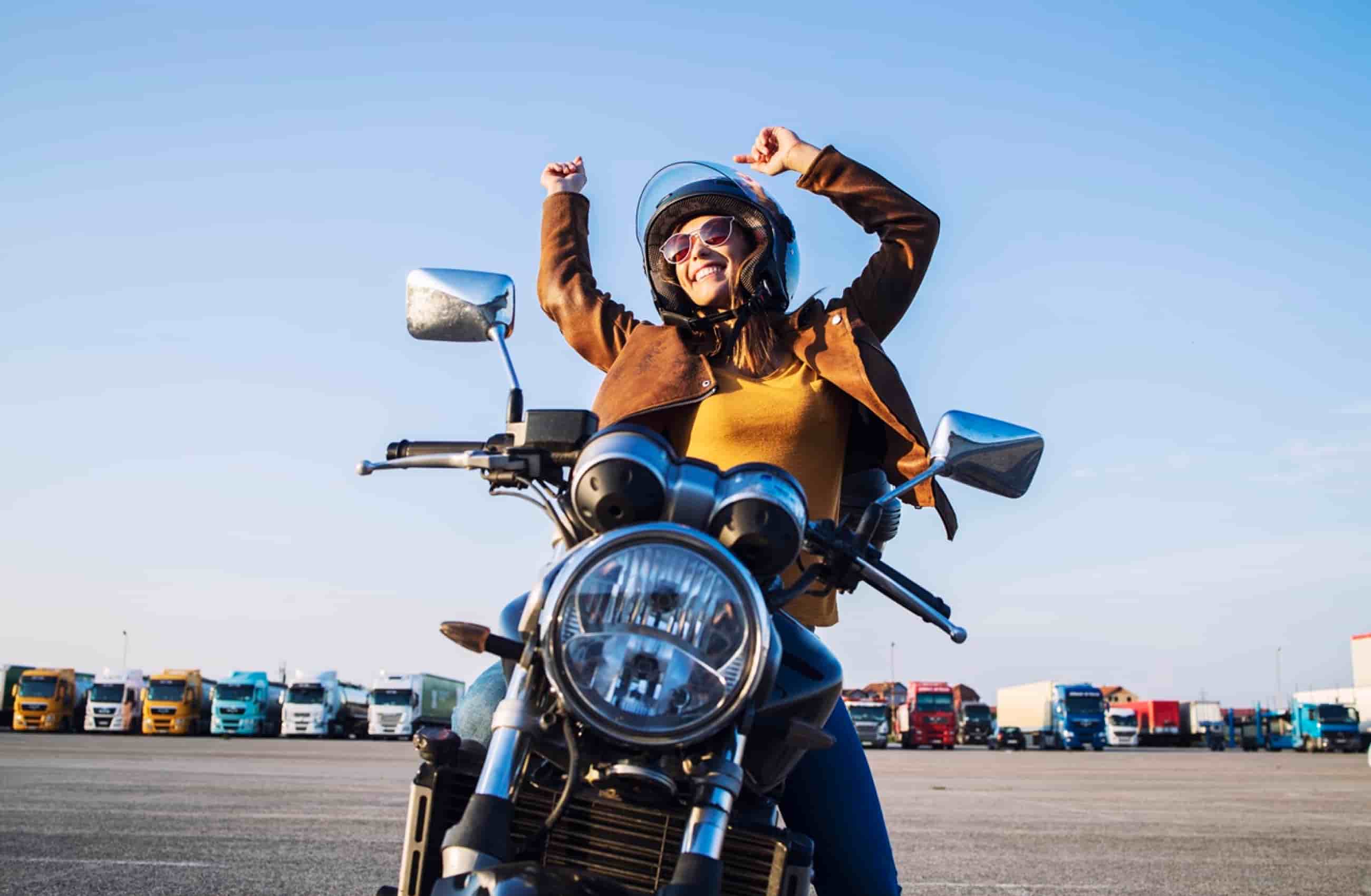 Mulher com jaqueta de couro marrom, óculos escuros e capacete. Ela está sentada em sua moto, sorrindo e com os braços pra cima, mostrando comemoração. Ao fundo vemos a imagem de vários veículos. A imagem representa como comprar uma moto parcelada.