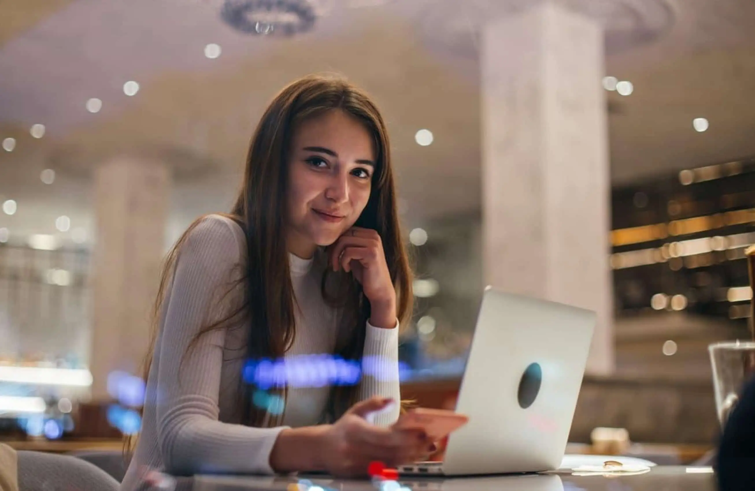 Uma mulher sorrindo sentada na mesa de um café com um notebook e um smartphone.&nbsp;
