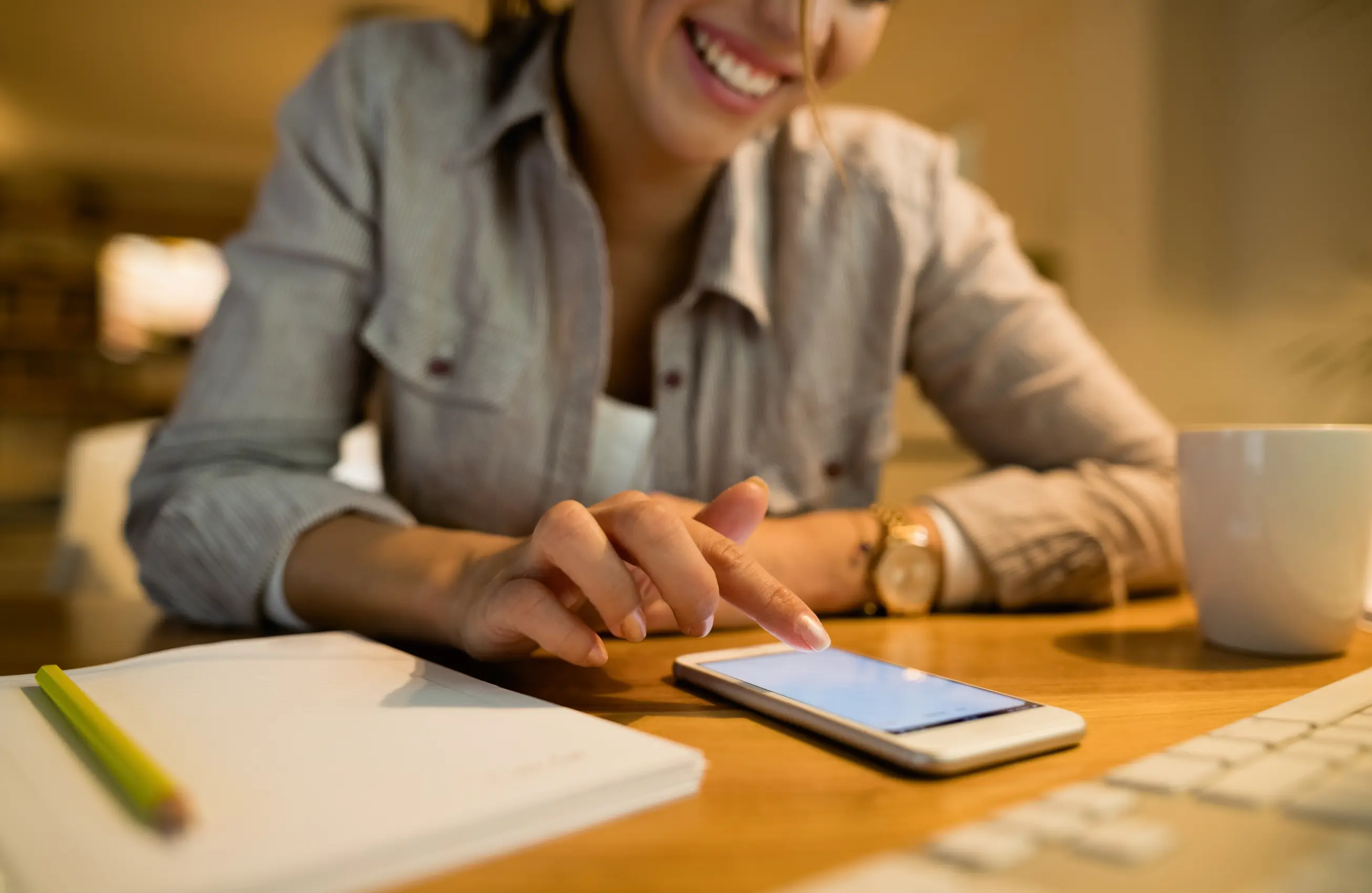 Mulher mexe na tela de um smartphone em uma mesa de escritório, com caderno, lápis e xícara de café ao redor; iluminação quente.
