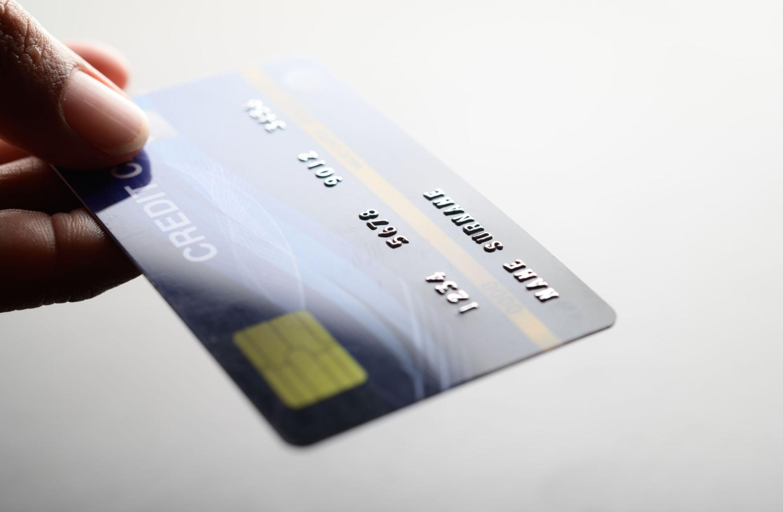 Mão segurando um cartão de crédito próximo à câmera, com o cartão parcialmente girado para mostrar o brilho das letras e números em relevo com fundo desfocado.