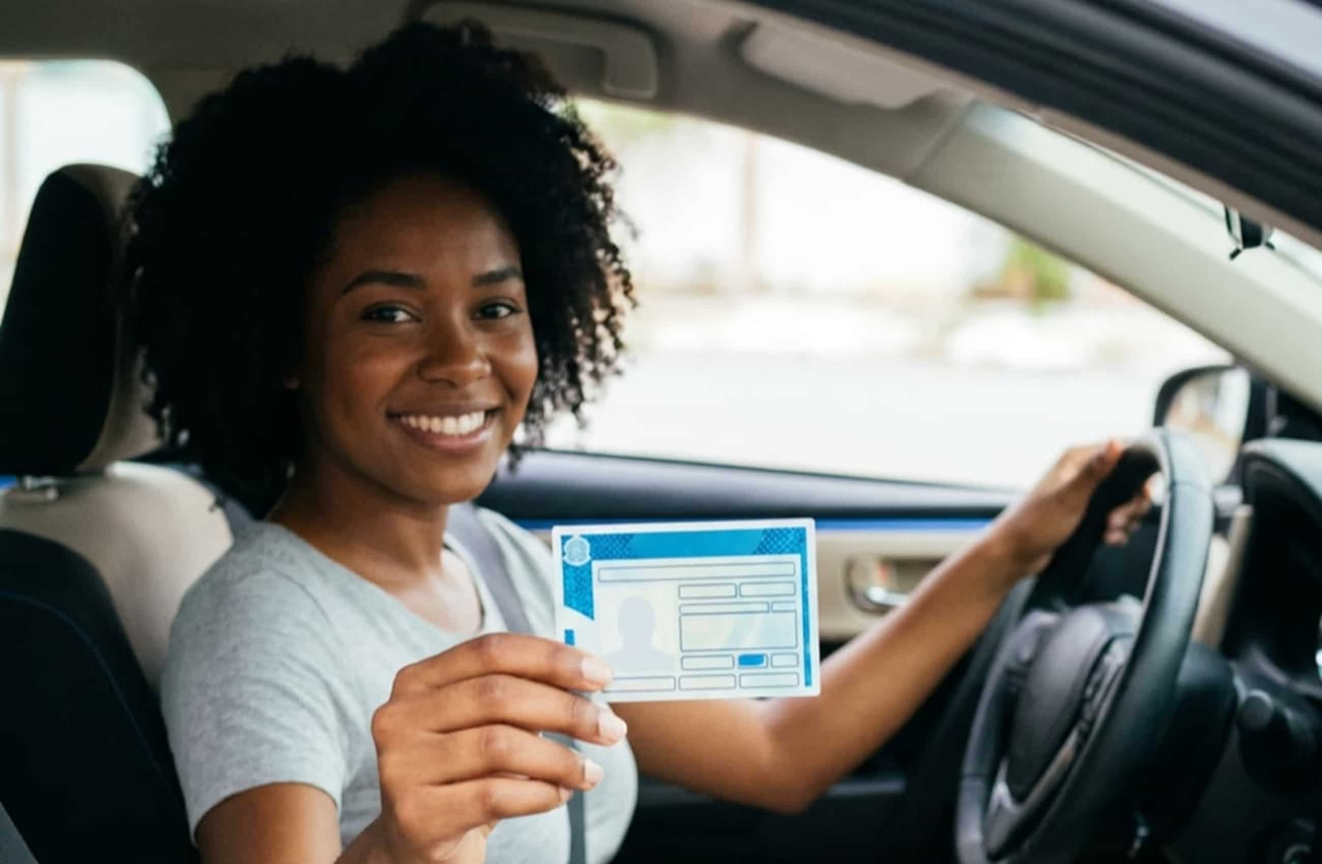 Mulher negra com camiseta cinza está sentada no banco do motorista de um carro. Ela está com uma das mãos no volante, e outra segurando uma CNH enquanto olha para a câmera.