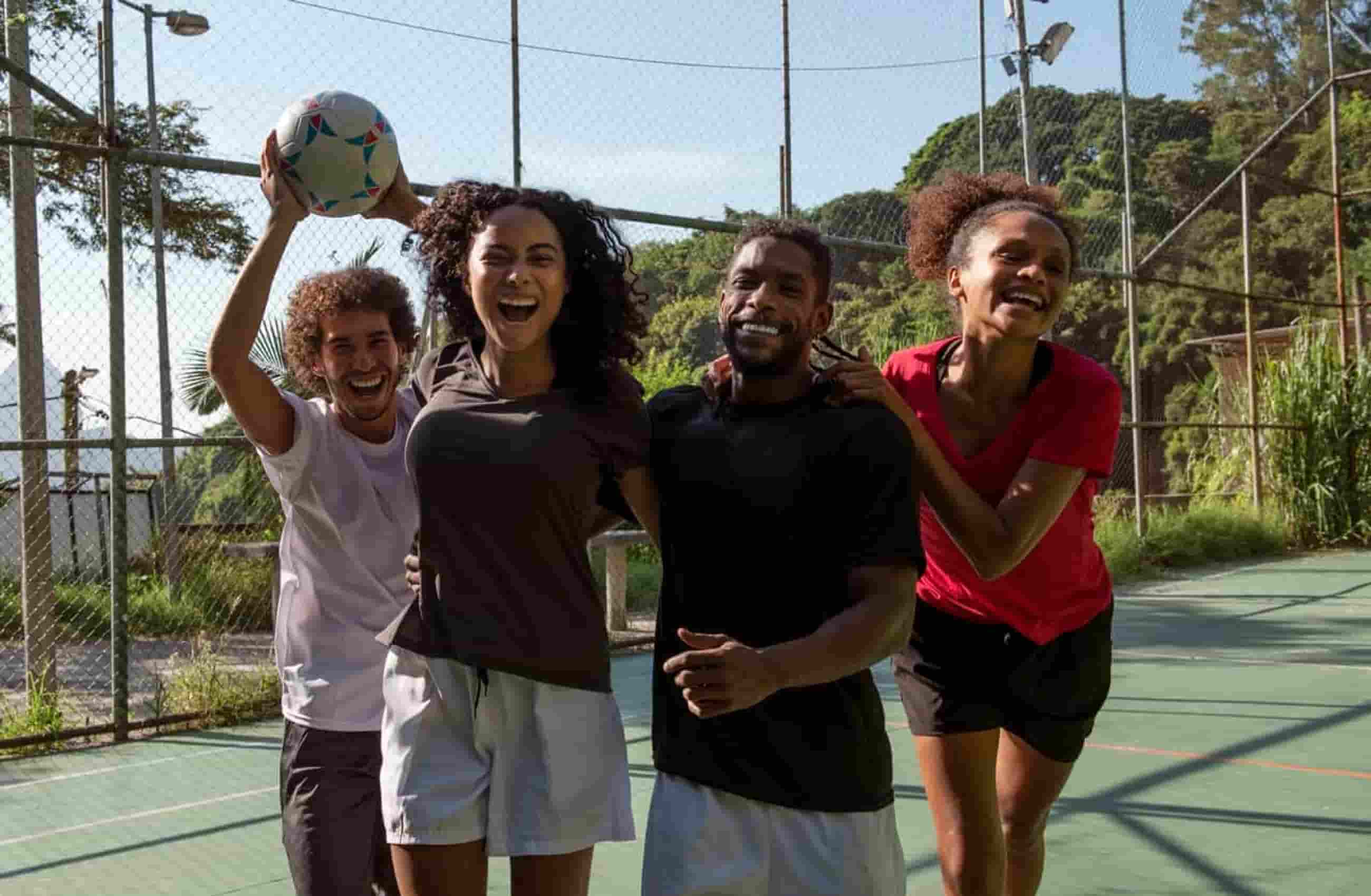 Quatro pessoas felizes sorrindo para a câmera enquanto praticam esportes. Elas ventem roupas esportivas e estão numa quadra.