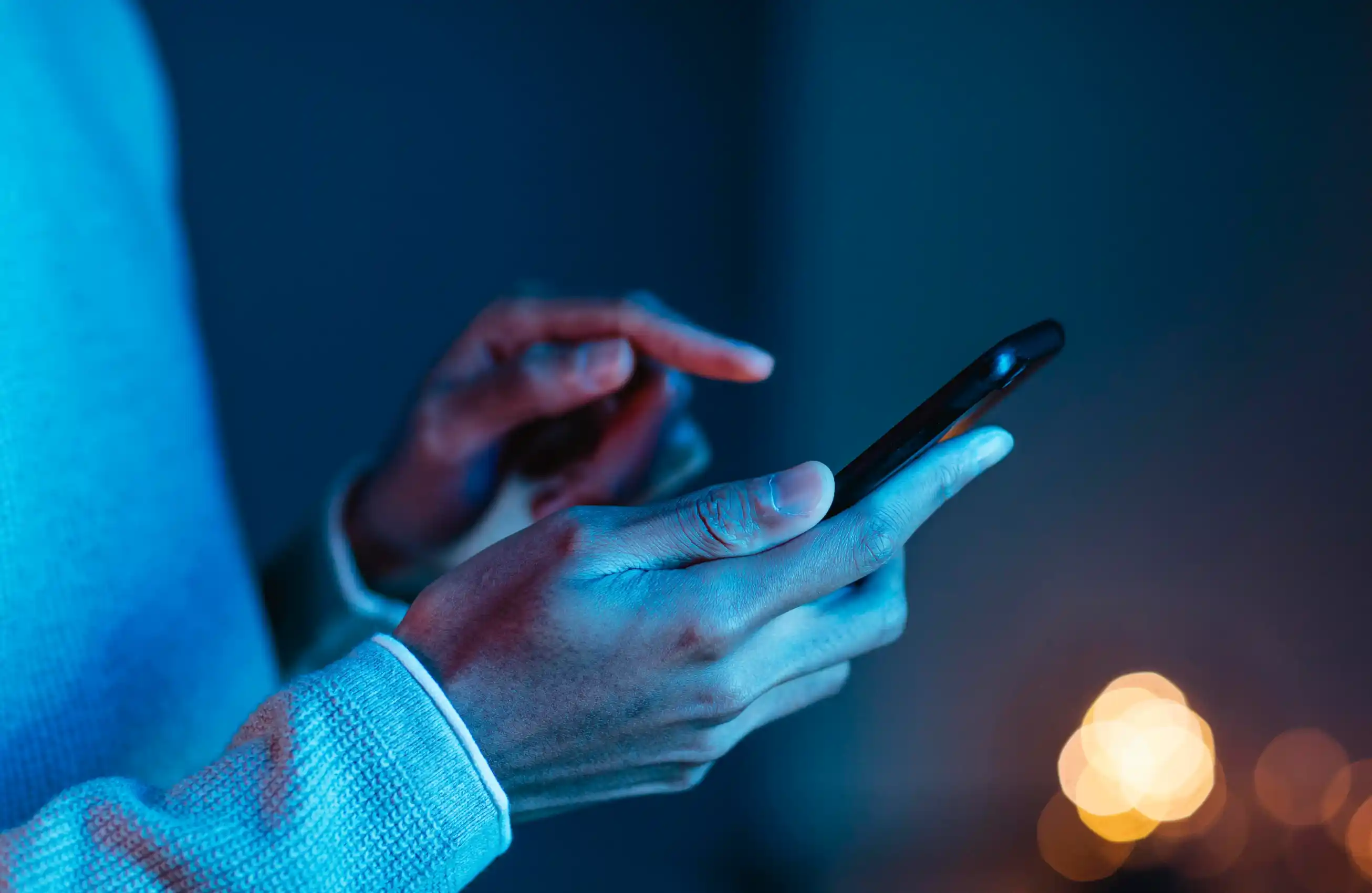 Imagem das mãos de um homem utilizando um smartphone. Ele utiliza um suéter e a iluminação do local é azul.