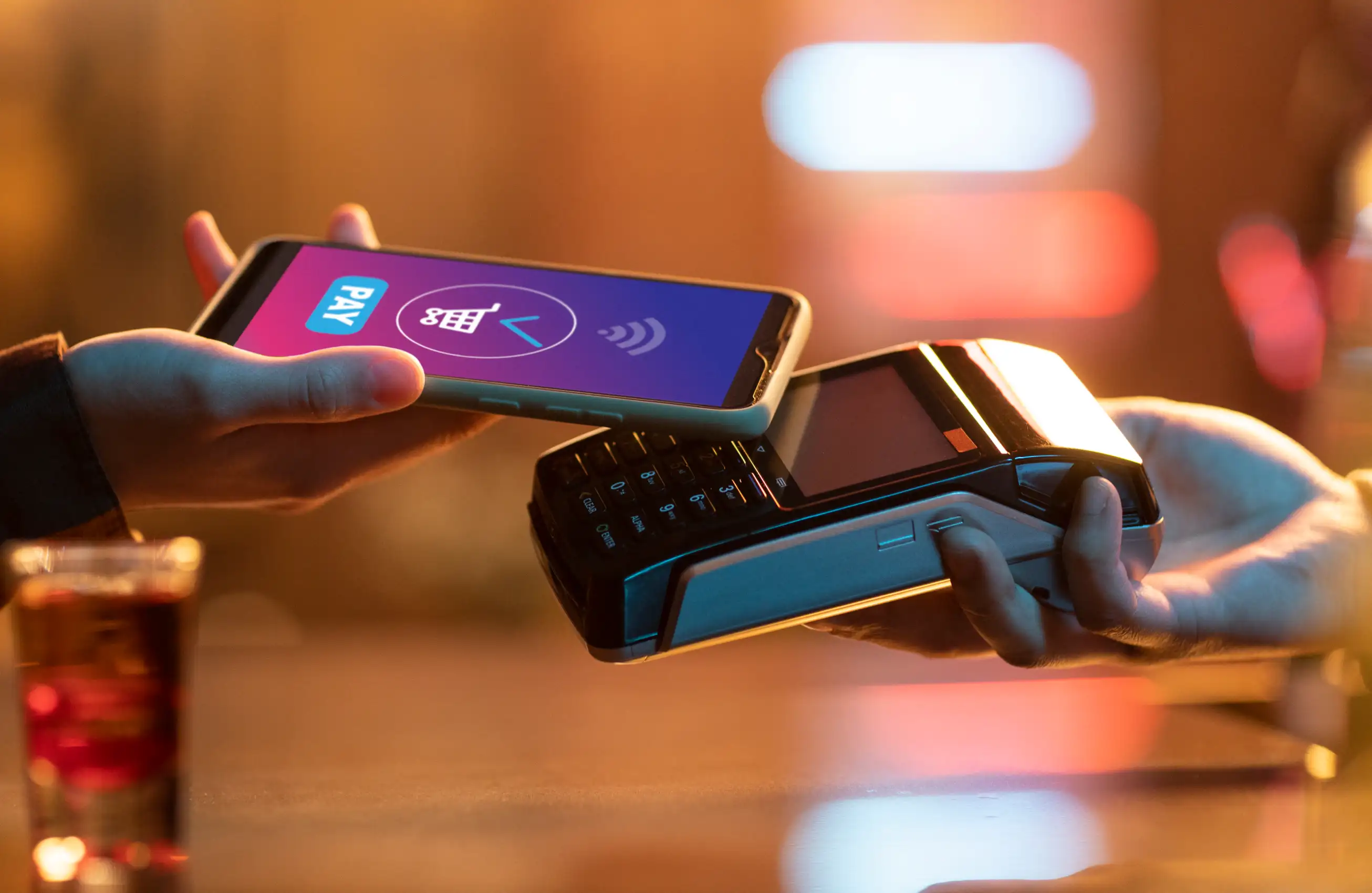 Pessoa segurando uma maquininha de pagamento enquanto outra aproxima o celular para realizar um pagamento por aproximação, foco nas mãos e nos dispositivos.