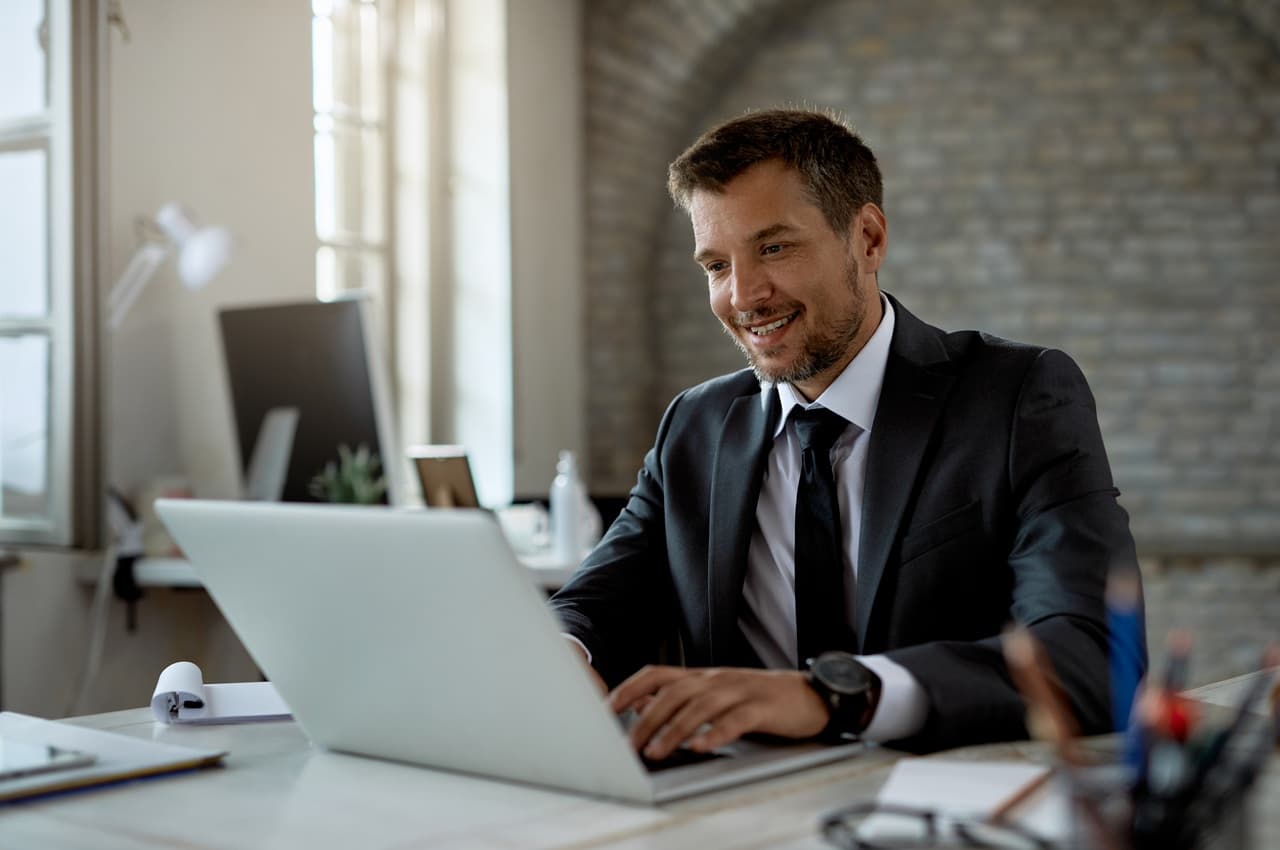 Homem sorridente, de terno e gravata, digitando em um notebook em uma mesa de escritório. Ele está entrando no Internet Banking PJ BV.