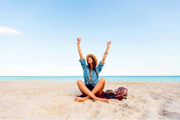 Mulher jovem com chapéu de palha sentada na areia de uma praia tropical, celebrando os feriados prolongados 2026 com os braços erguidos em sinal de liberdade. O cenário exibe um céu limpo e mar azul ao fundo.