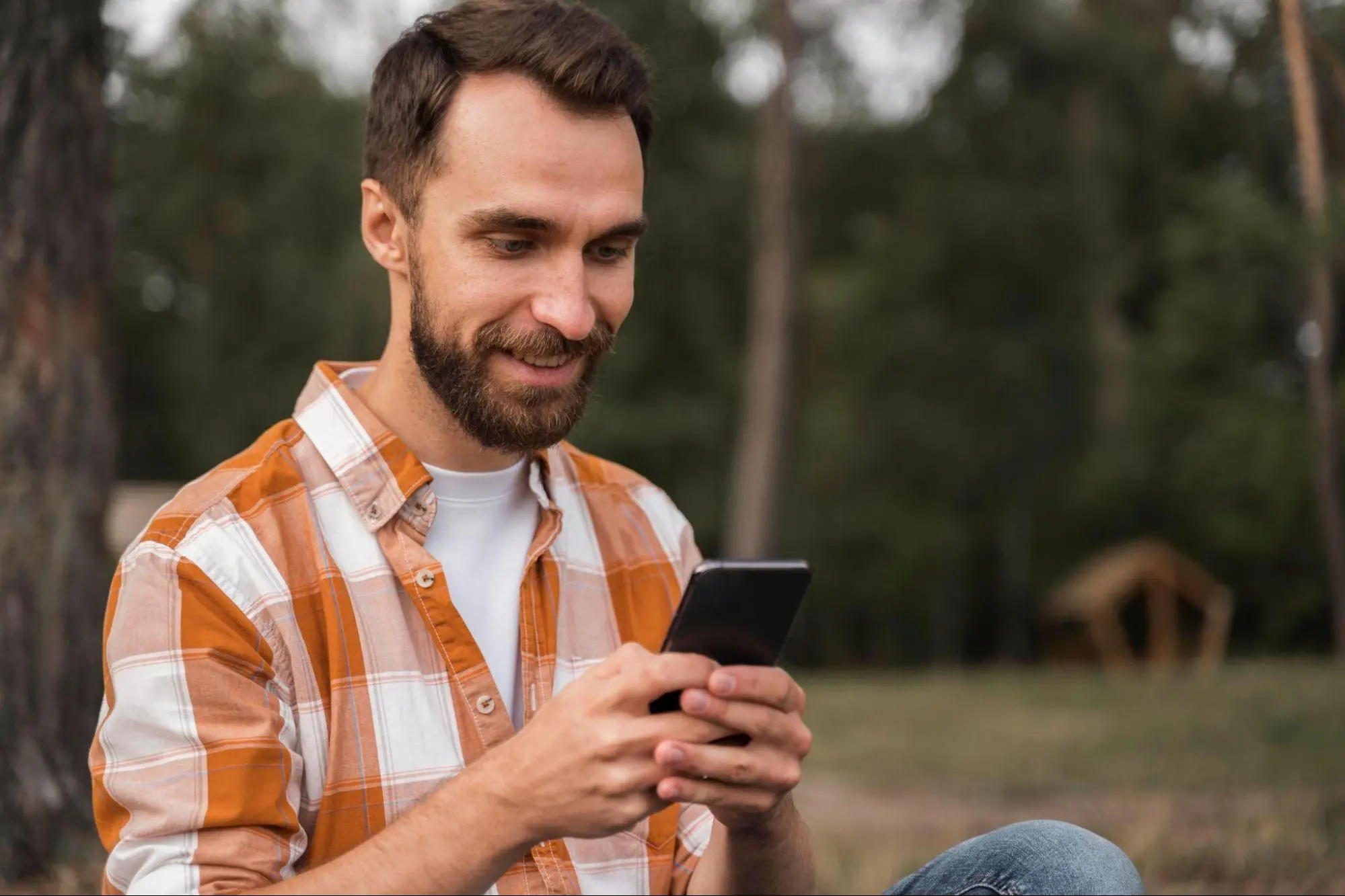 Homem sorridente com barba, usando uma camisa xadrez laranja, segurando um smartphone. Ele está ao ar livre, próximo a árvores.