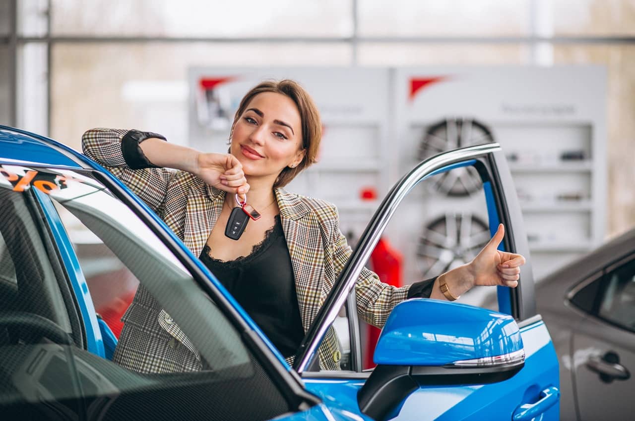 Mulher segurando as chaves de um carro em uma das mãos e sorriso no rosto, ao lado de um veículo azul com a porta do motorista aberta. Ela está em uma concessionária de automóveis.