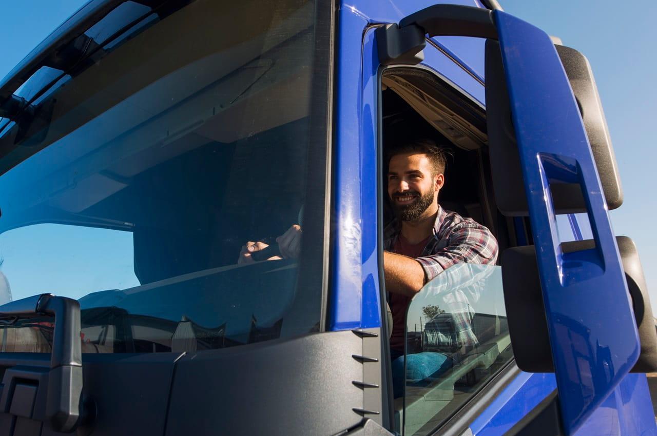 Homem com barba sorrindo e dirigindo um caminhão com detalhes na cor azul, com um céu claro ao fundo.