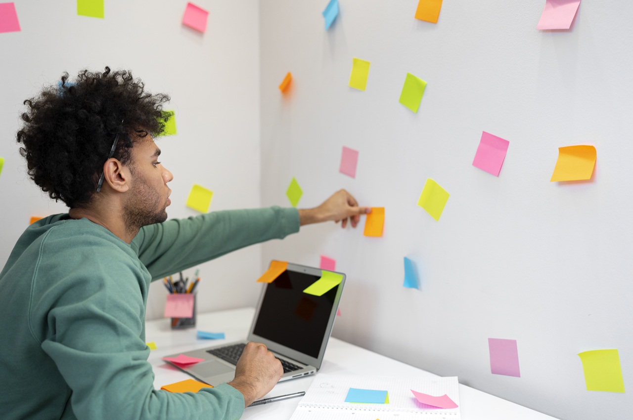Homem jovem colando notas adesivas em uma parede branca coberta de post-its coloridos. Ele está sentado em frente a um laptop e montando o seu projeto pessoal.
