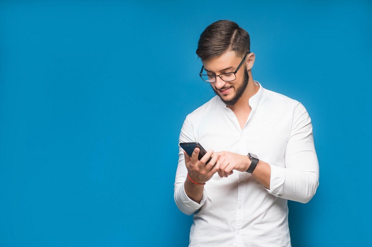 Homem sorridente vestindo blusa social branca, óculos de grau e relógio. Ele acessa FGTS Completão BV pelo seu celular. O fundo da imagem é azul.
