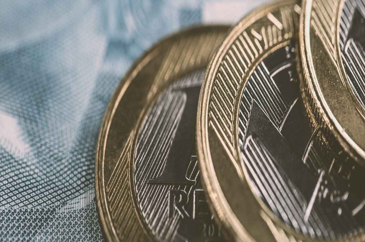 Foco em moedas de um real (R$1) em cima de notas de cem reais (R$100,00).