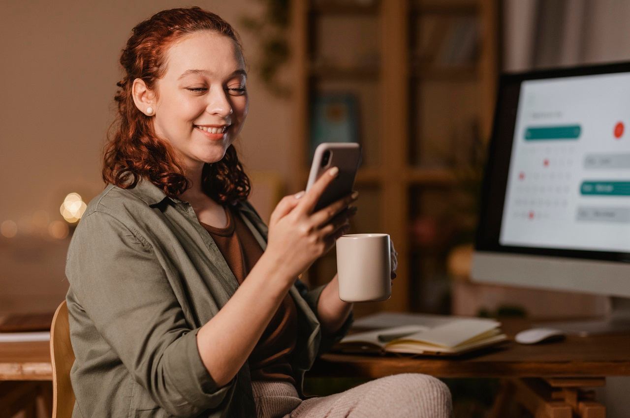 Mulher sorridente de cabelos ruivos médio, segurando xícara de café e acessando empréstimo FGTS pelo celular. Ela veste blusa marrom básica, calça cinza de moletom e está sentada em frente a um computador.