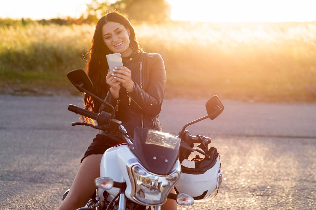 Mulher sorridente de jaqueta de couro usando um smartphone enquanto está sentada em uma motocicleta branca durante o pôr do sol.