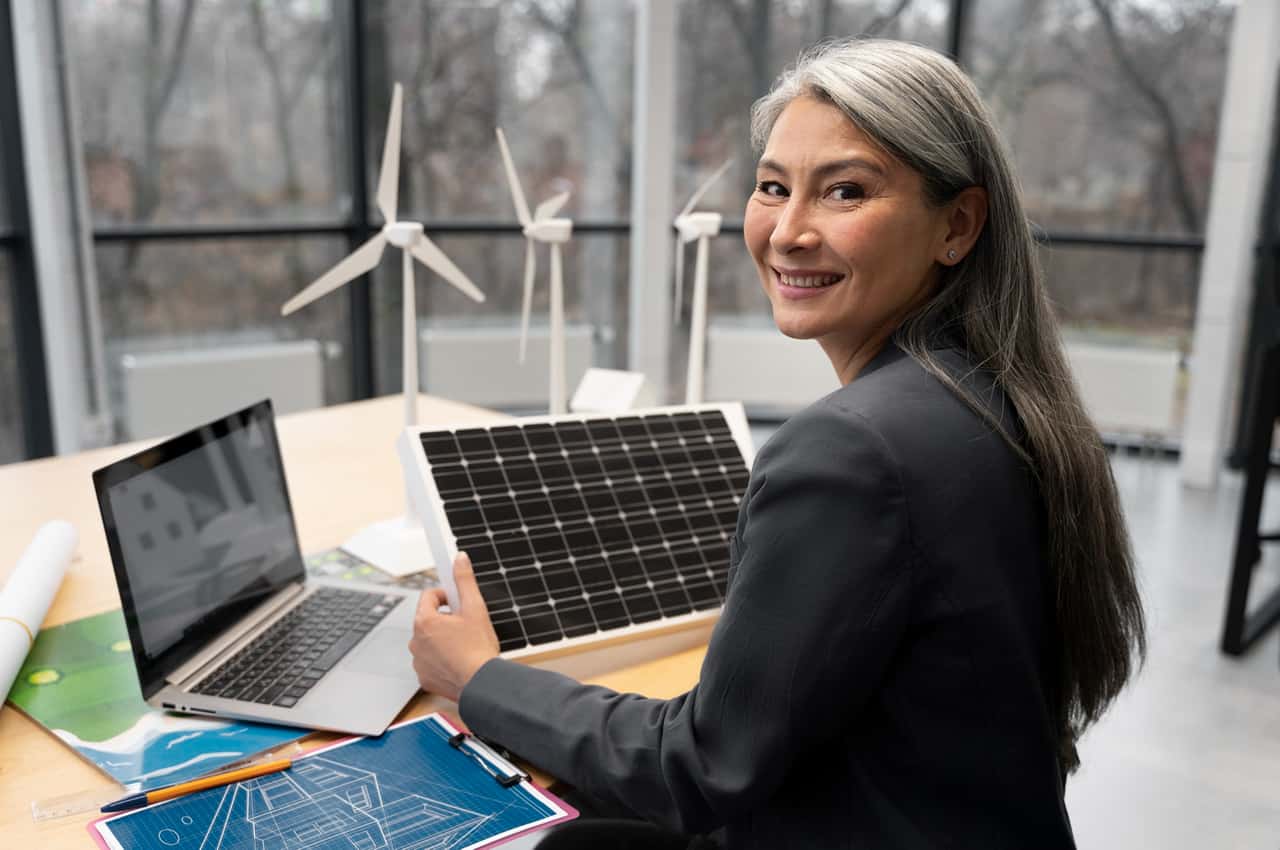 Engenheira sorrindo enquanto segura painel solar em um escritório, com laptop e maquetes de turbinas eólicas.