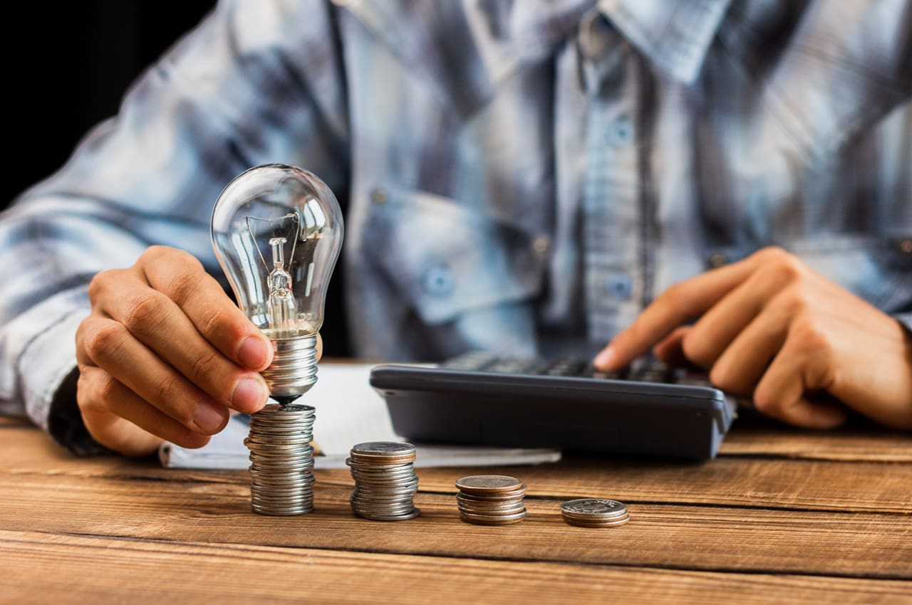 Pessoa segurando uma lâmpada sobre pilhas de moedas, simbolizando como evitar desperdício de energia elétrica.