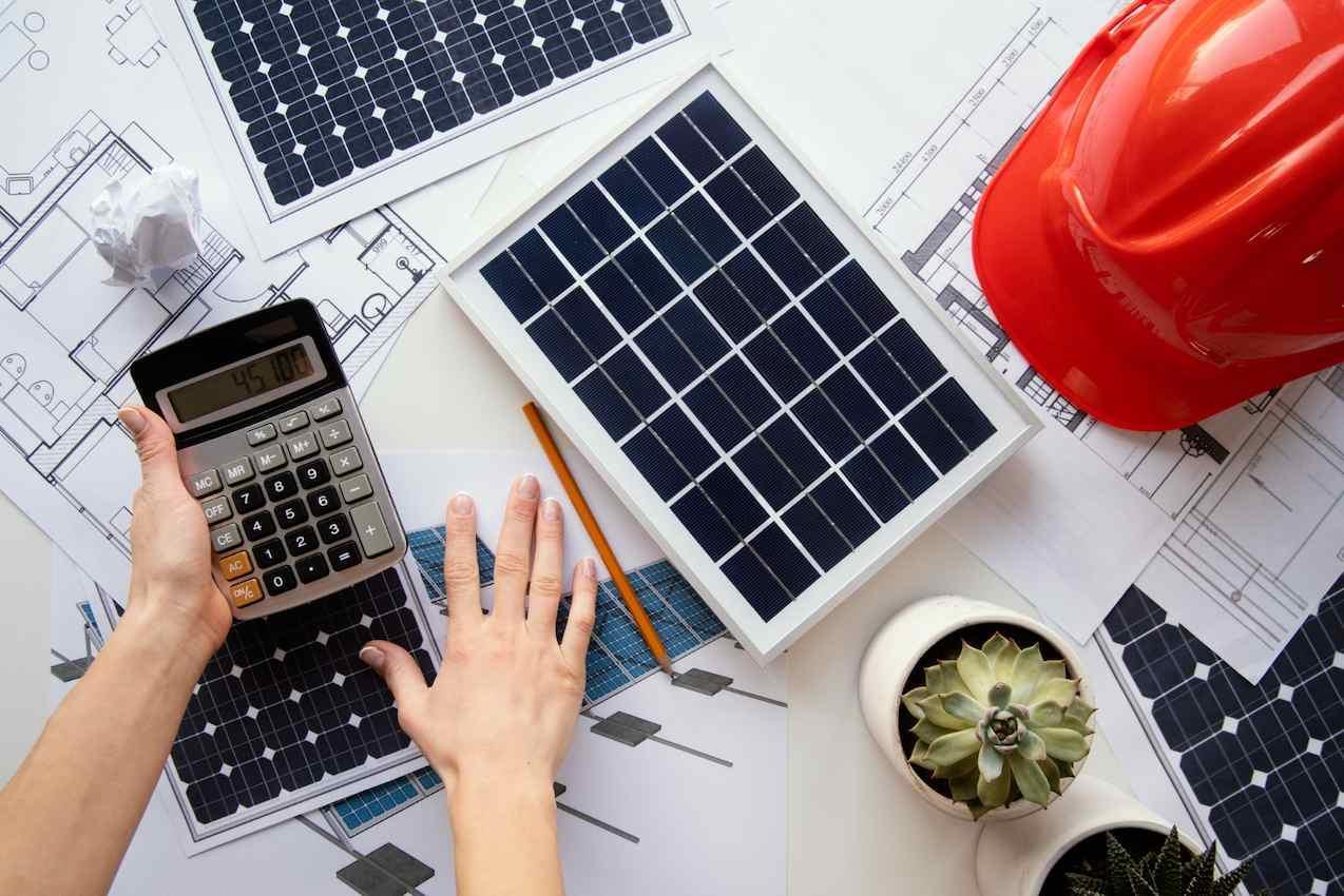Mesa com projetos de painéis solares, uma calculadora, uma planta suculenta e um capacete vermelho, simbolizando a transferência de créditos de energia solar.
