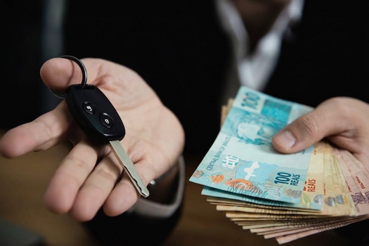 Close-up de uma pessoa de terno estendendo a chave de um carro em uma mão e um maço de notas de reais na outra.
