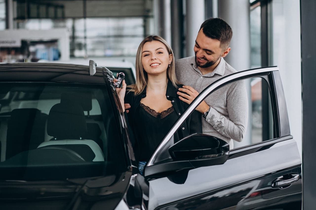 Casal sorridente ao lado de um carro preto com a porta aberta, em uma concessionária. A mulher segura a chave do veículo.