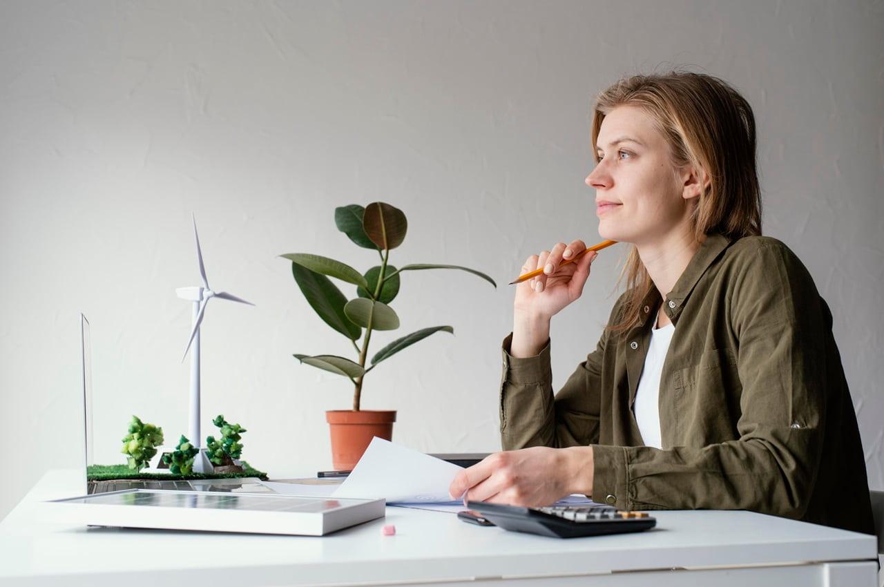 Mulher pensativa e esboçando um sorriso, com plantas, modelos de energia renovável e símbolo de sustentabilidade na mesa. Ela está refletindo sobre a economia de baixo carbono.