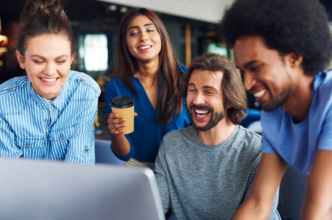 Grupo diverso de quatro colegas de trabalho sorrindo enquanto olham juntos para a tela de um computador, em celebração à obtenção do selo GPTW.