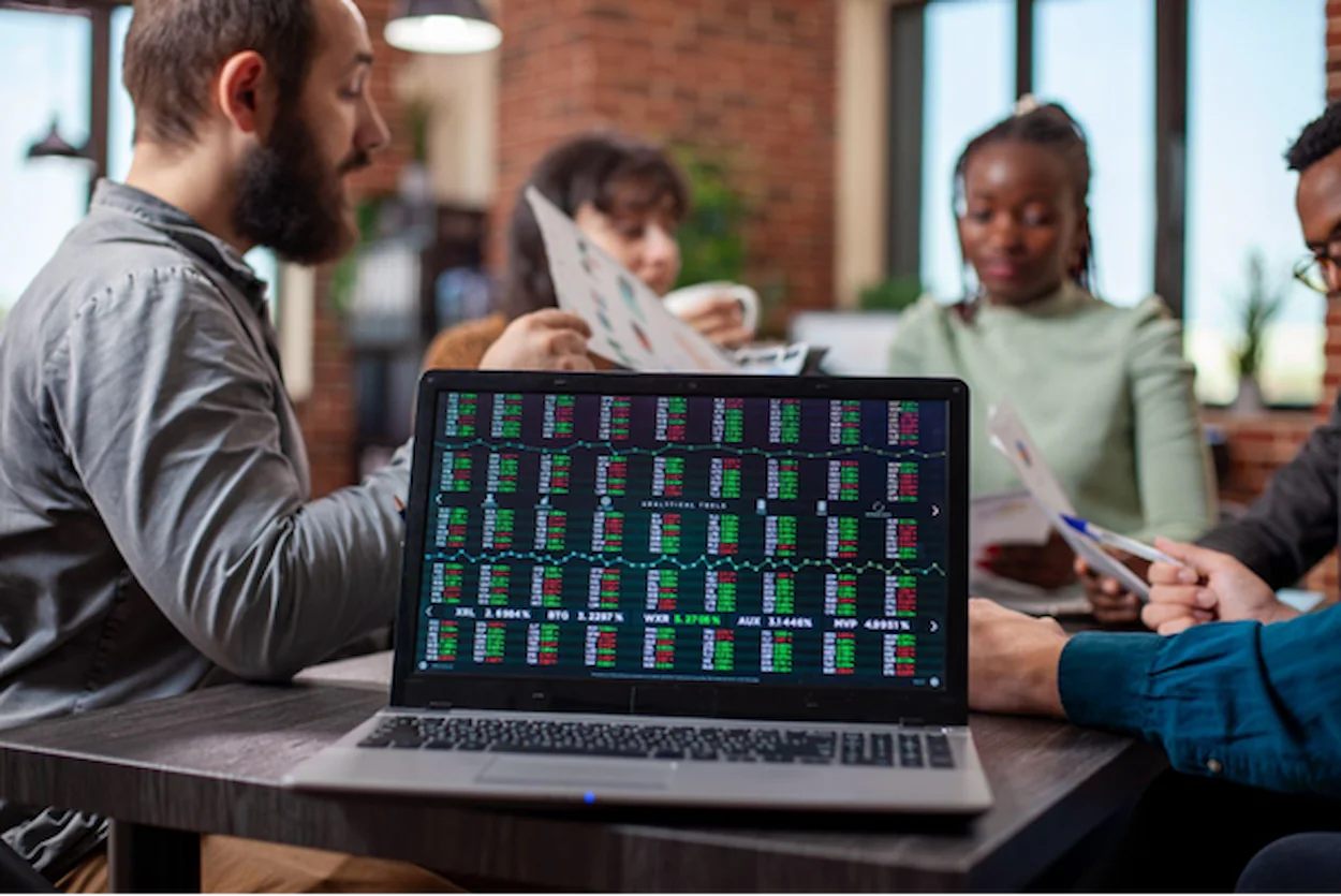 Um notebook com gráficos e dados financeiros está em evidência. Ao fundo, pessoas sentadas em uma mesa analisam os dados em papéis.