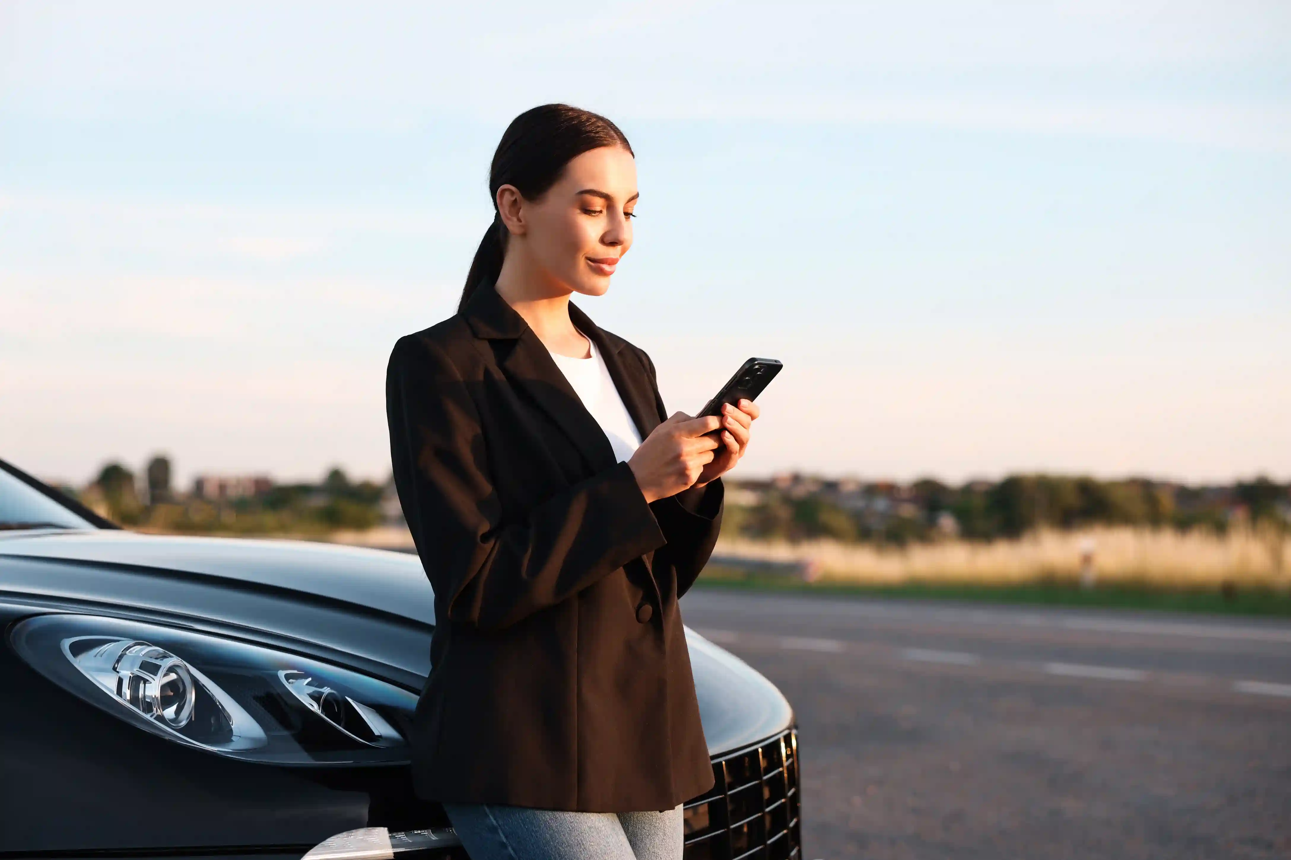 Mulher ao lado do carro consultando o celular na estrada, ilustrando a contratação e assistência do Seguro RCF-V.&nbsp;
