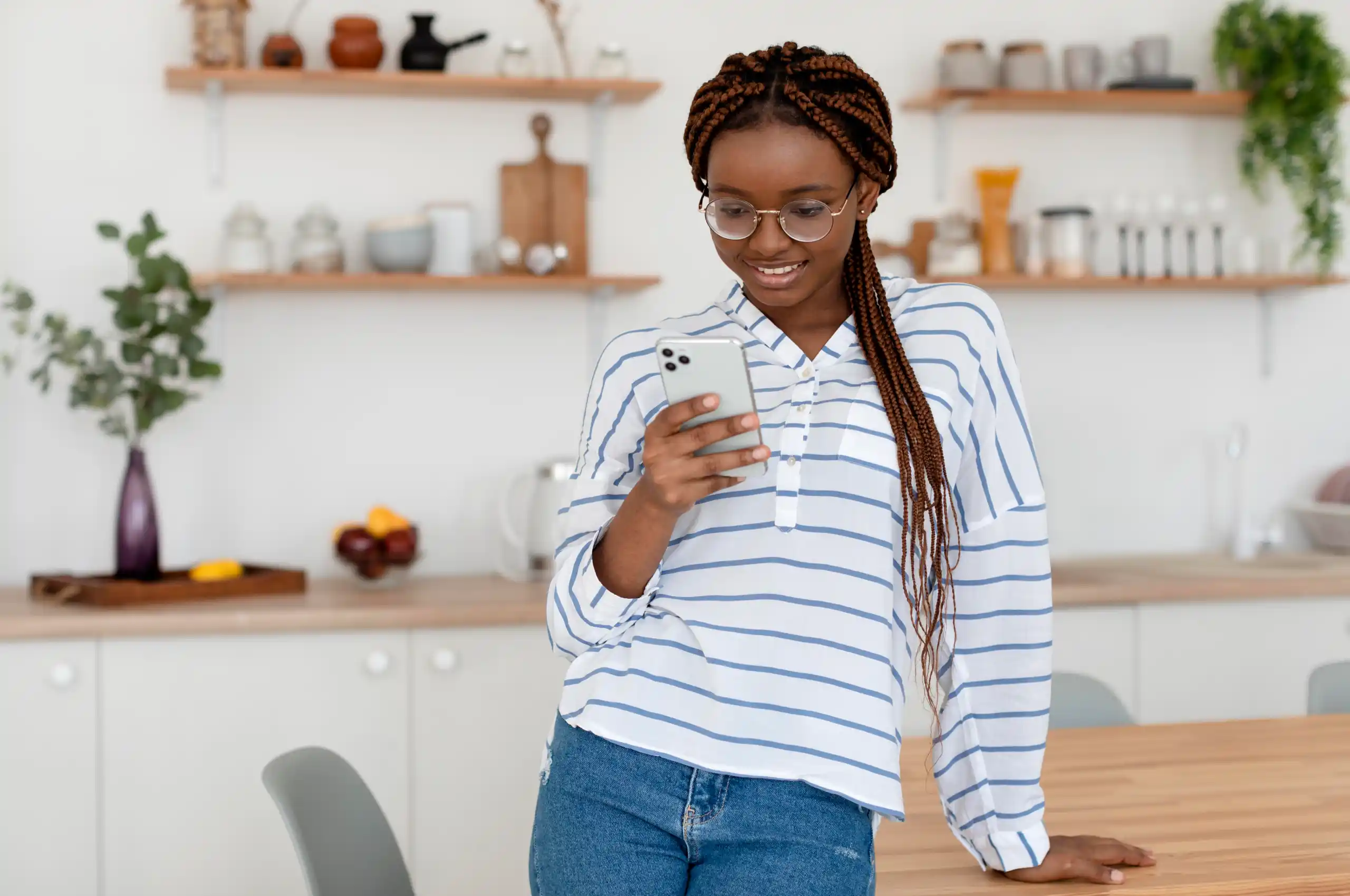 Uma mulher apoiada na mesa de estar de uma cozinha, enquanto utiliza um smartphone e sorri.