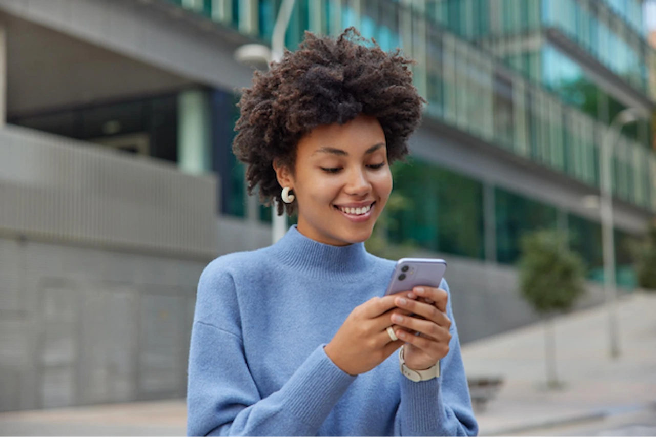 Mulher de suéter azul sorri enquanto usa o celular na rua, fazendo uma recarga de celular pelo aplicativo.