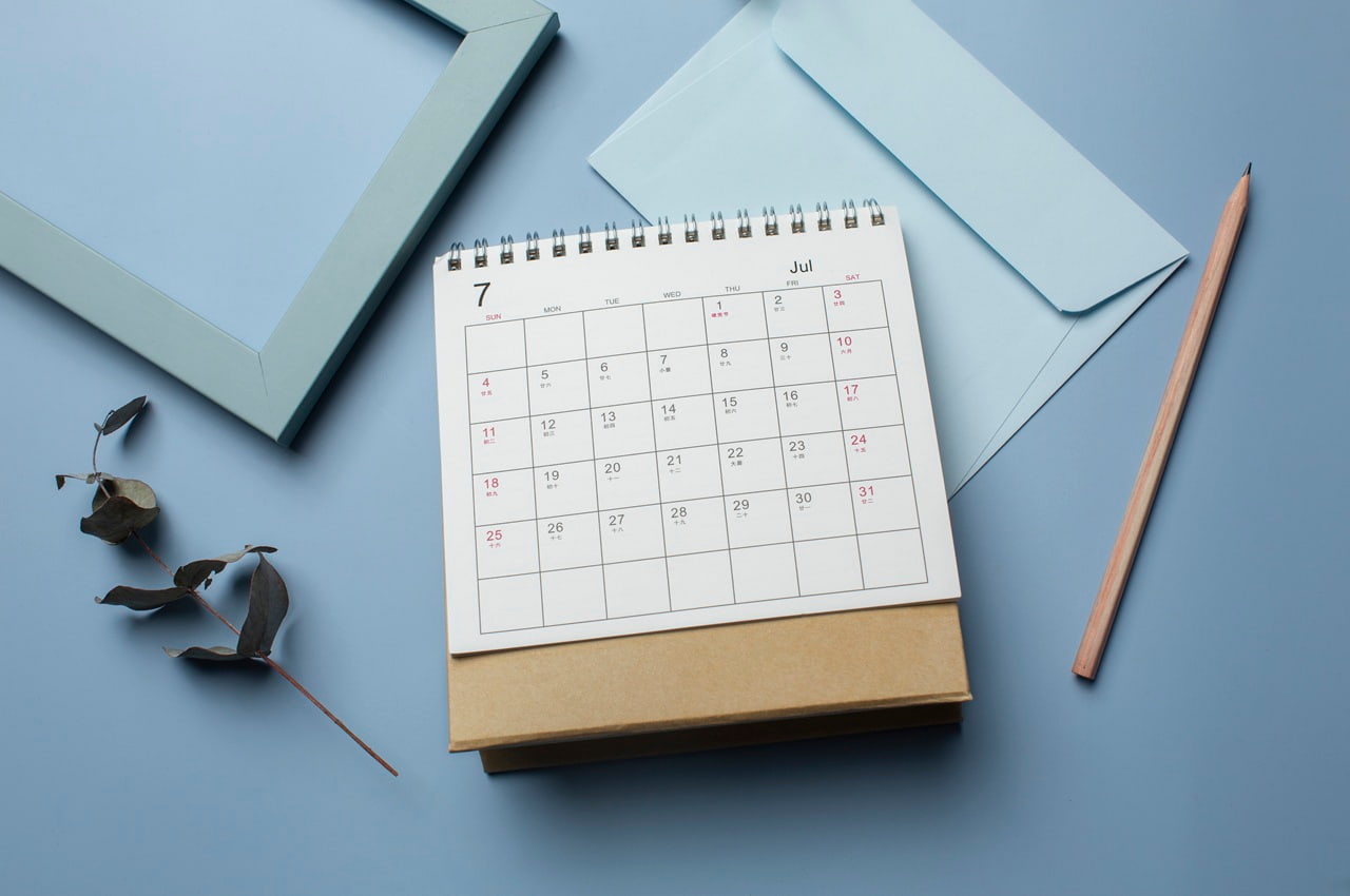Vista superior de um calendário de mesa, um envelope, um lápis e uma moldura organizados sobre um fundo azul.