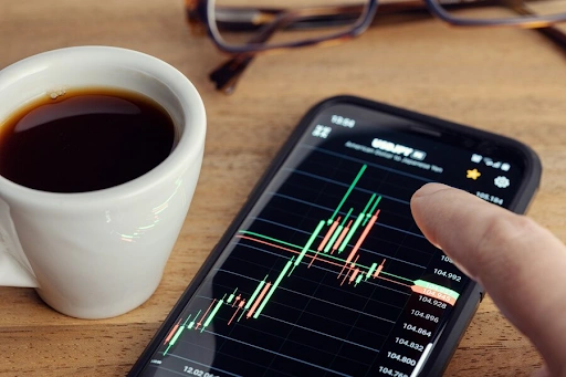 Xícara de café ao lado de um smartphone exibindo gráficos financeiros, com uma mão apontando para os dados na tela. Óculos estão visíveis ao fundo. 