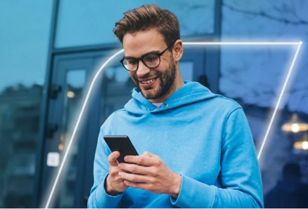 Homem de óculos e moletom azul sorri ao usar o celular. Ao fundo, prédio espelhado e uma linha geométrica brilhante em neon.