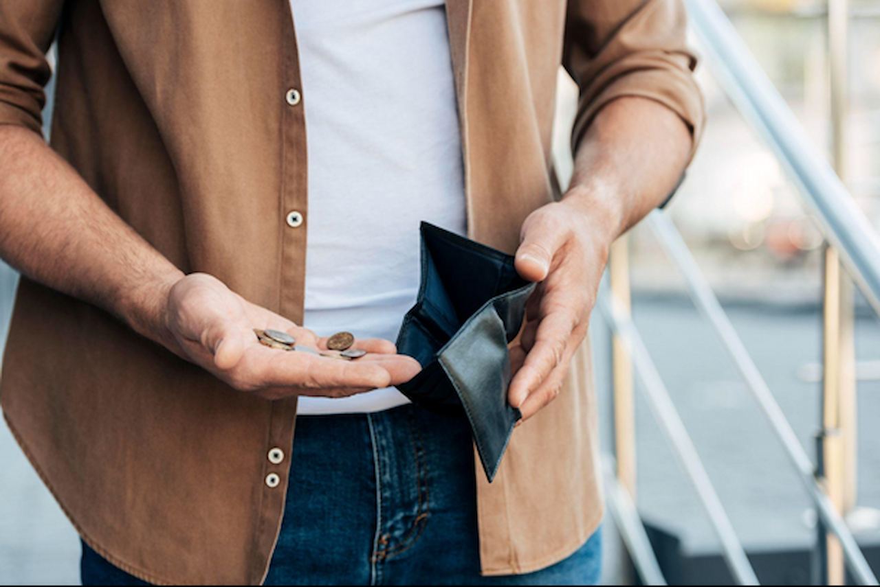 Pessoa segurando moedas na palma da mão esquerda e uma carteira aberta na mão direita. Foco no dinheiro visível, com a pessoa vestindo camisa marrom e jeans, em cenário urbano.
