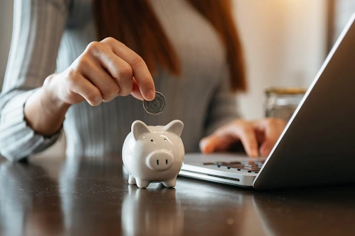 Mulher colocando moeda em cofre porquinho em mesa com notebook, representando economia ou investimento.