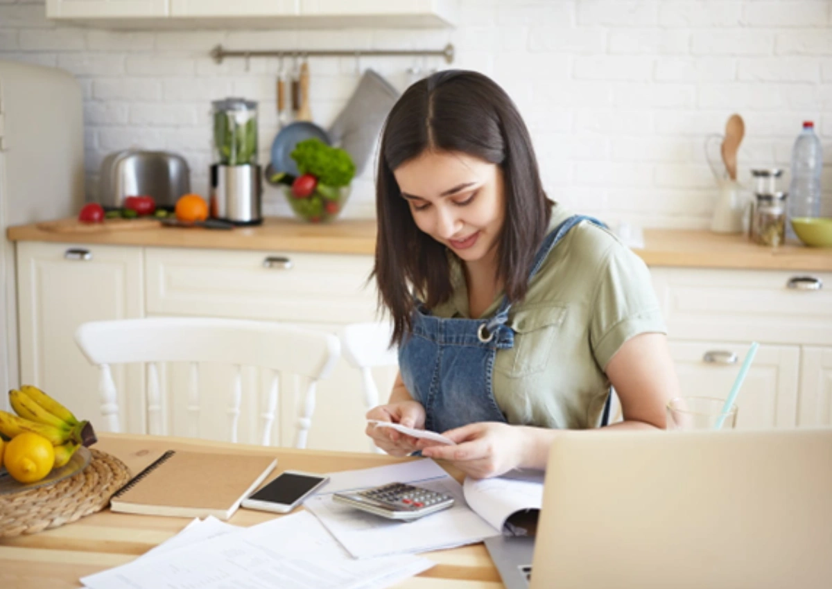 Mulher em mesa de cozinha. Há papéis espalhados, uma calculadora e celular na mesa, ao lado de uma fruteira. A mulher verifica no papel como funciona a liquidação antecipada no BV. Ao fundo, há uma bancada com materiais e utensílios de cozinha.