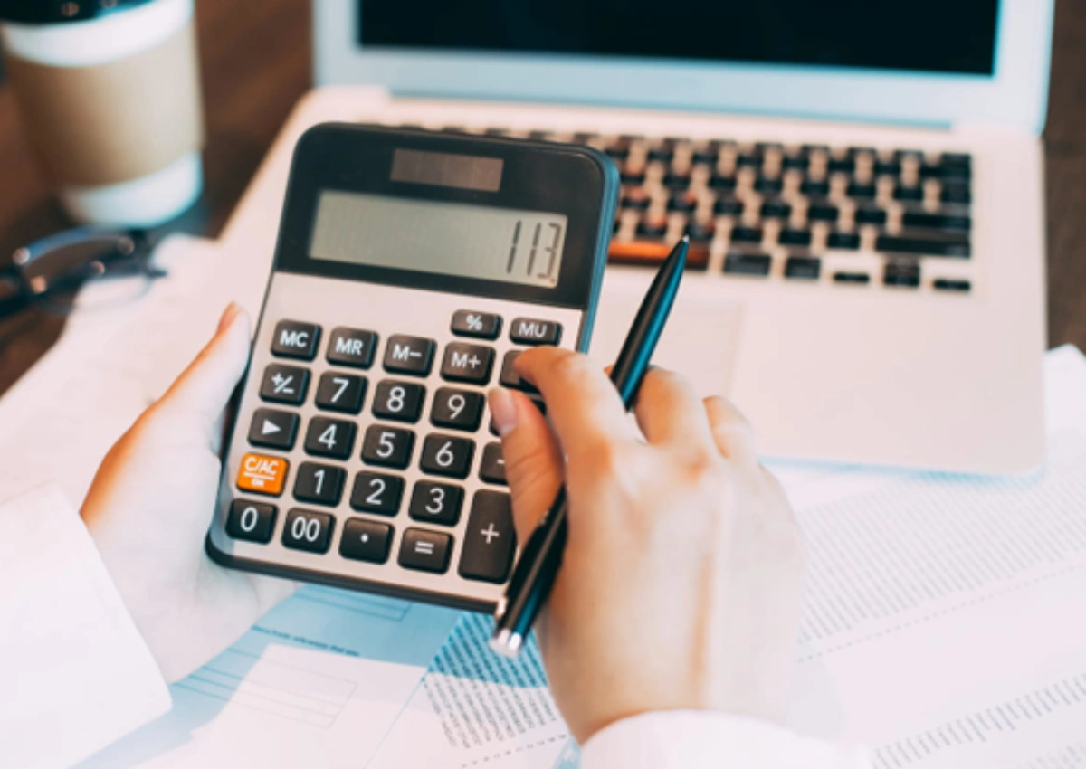 Duas mãos segurando uma calculadora e uma caneta, apoiadas sobre papéis. 