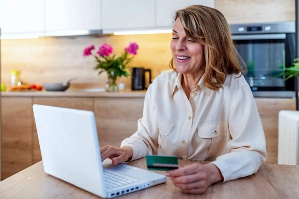 Mulher sentada em frente à mesa, em ambiente de cozinha. Está esquisando sobre antecipação de parcelas em um laptop com um cartão na mão. 