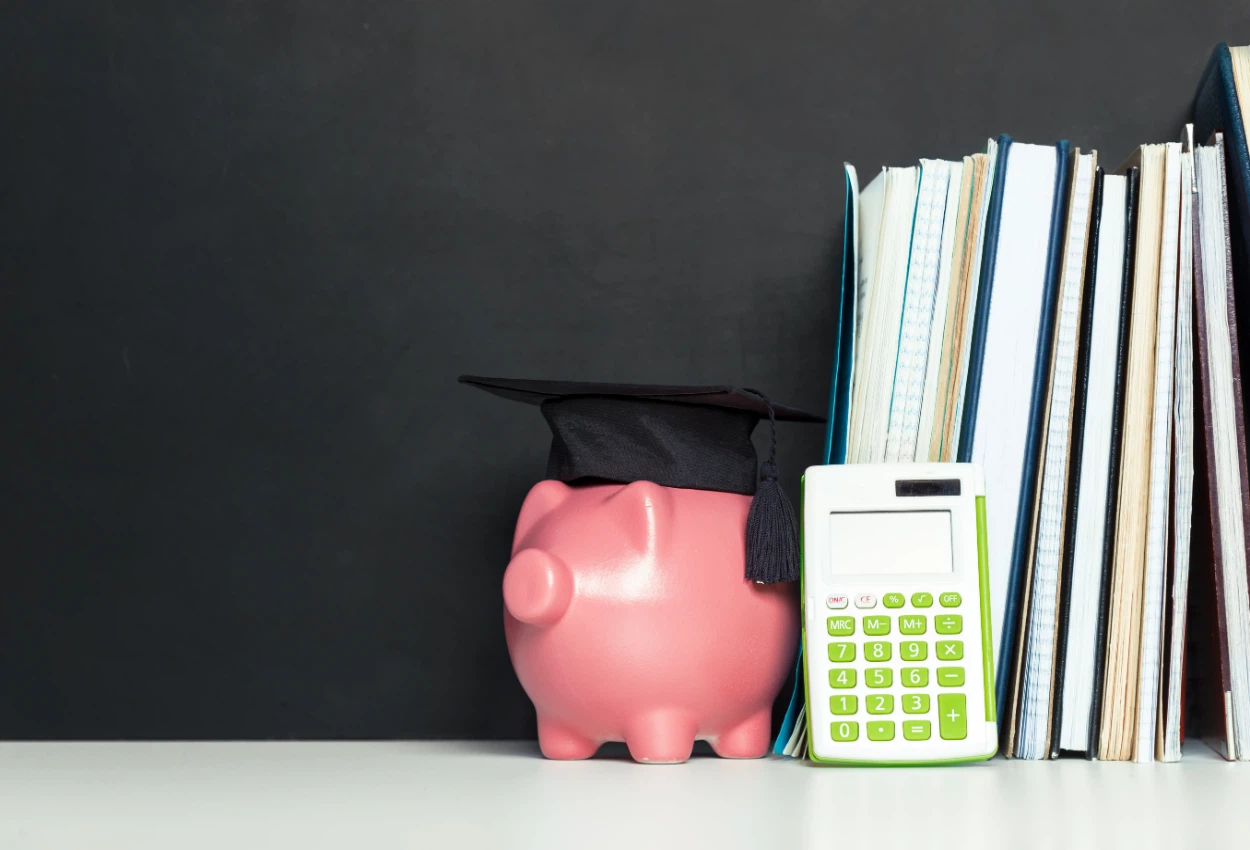 Cofrinho de porco com chapéu de formatura ao lado de livros e calculadora, simbolizando organização financeira para objetivos pessoais.