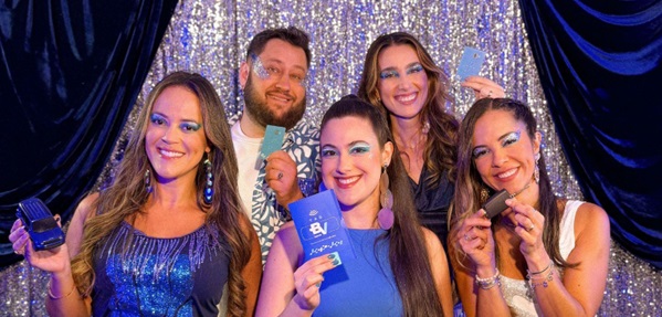 Grupo de foliões sorridentes em um cenário festivo de brilho, segurando cartões e a tag do Banco BV, representando a segurança no Carnaval 2026.