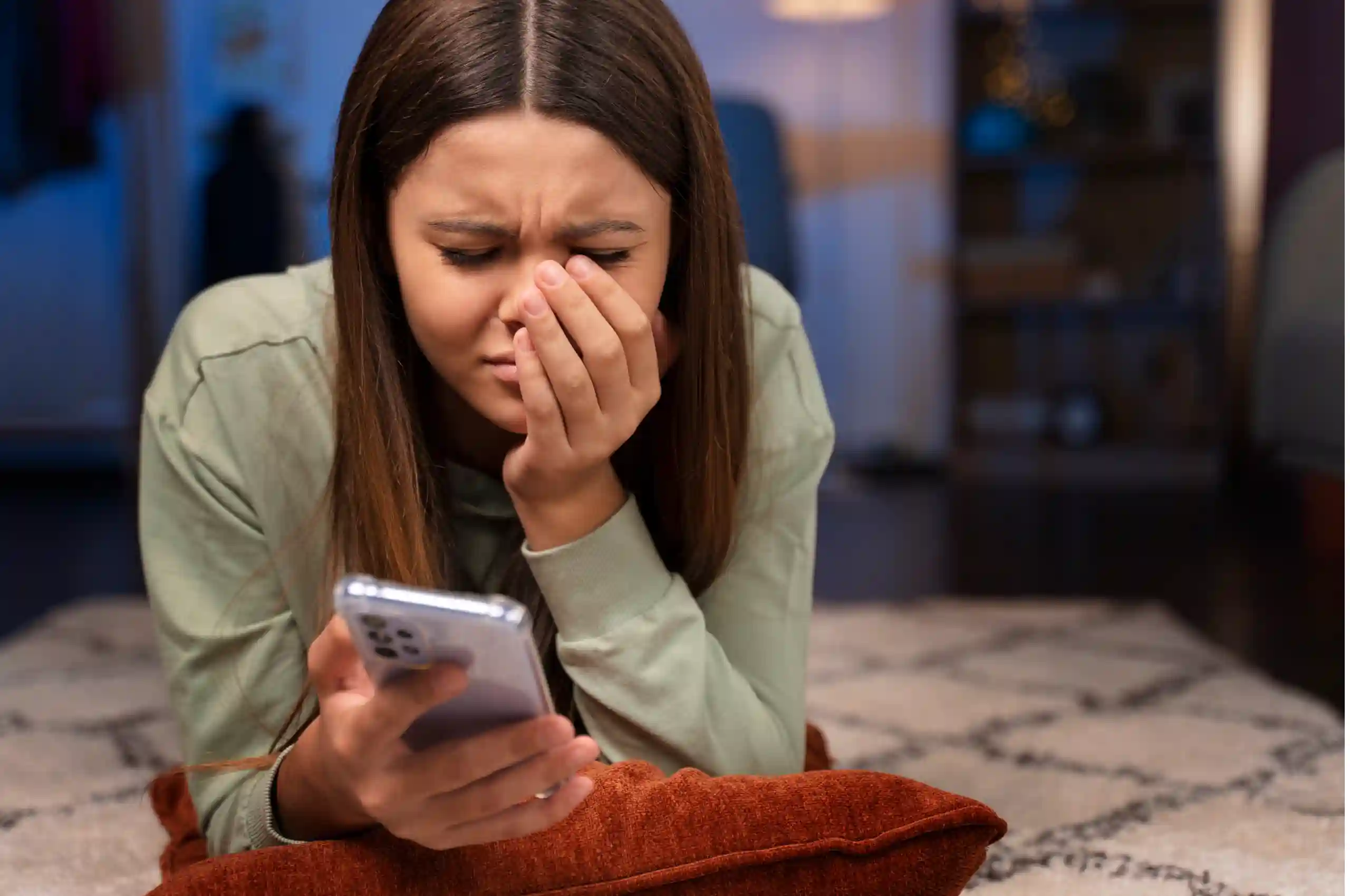 Uma mulher deitada sobre o chão de uma sala de estar. Ela segura um smartphone com a mão direita enquanto apoia a esquerda no rosto com uma expressão de procupação.