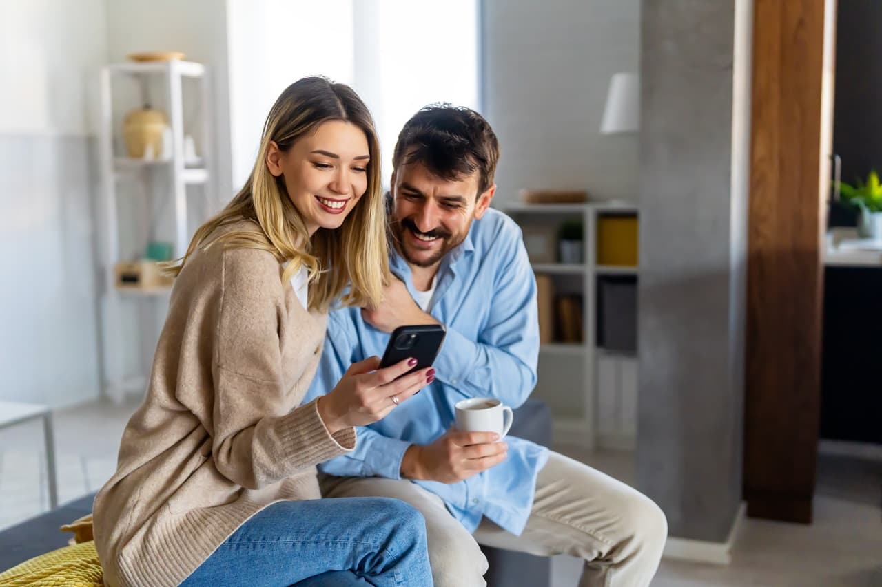 Casal em uma sala, sentado em um banco, olhando para celular na mão.