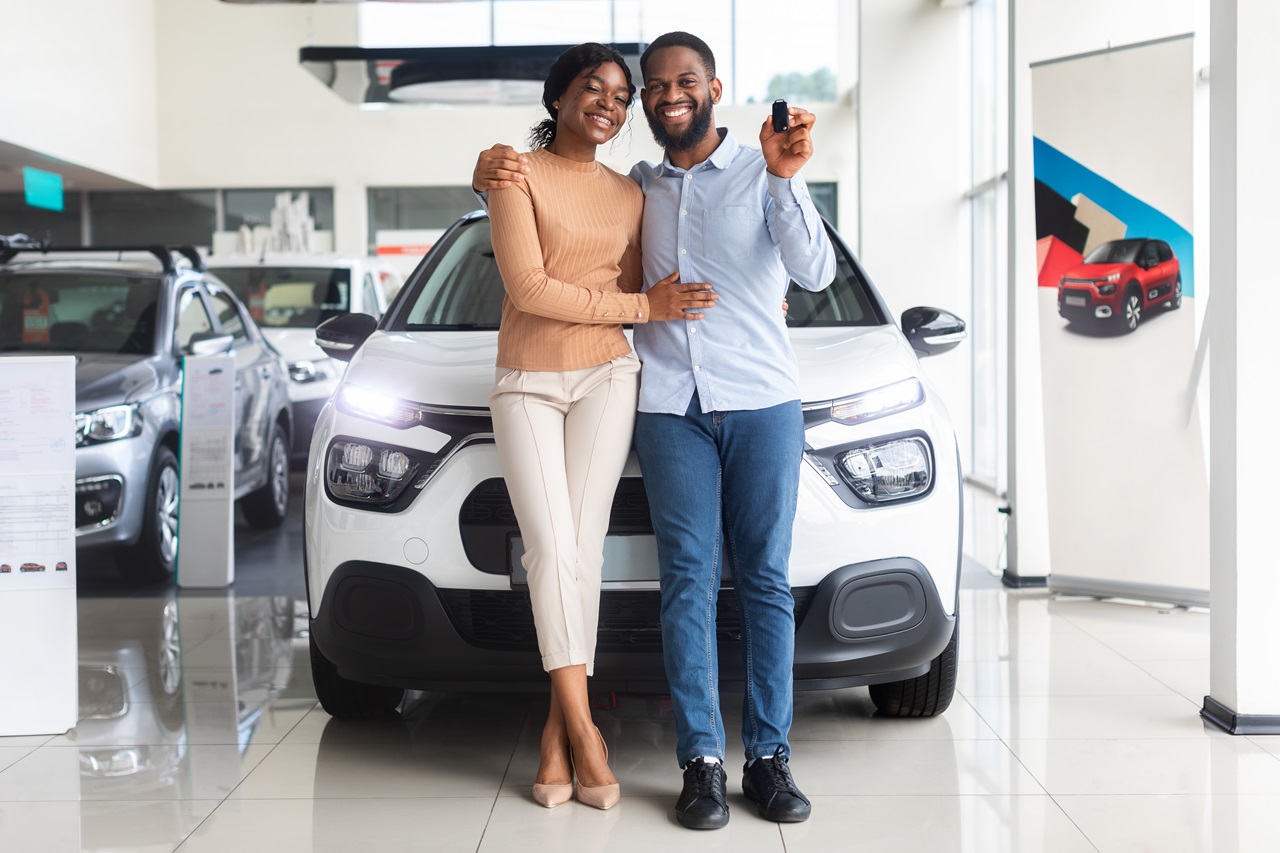 mulher e homem sorrindo em frente a um carro branco, o homem está segurando uma chave de carro na mão.