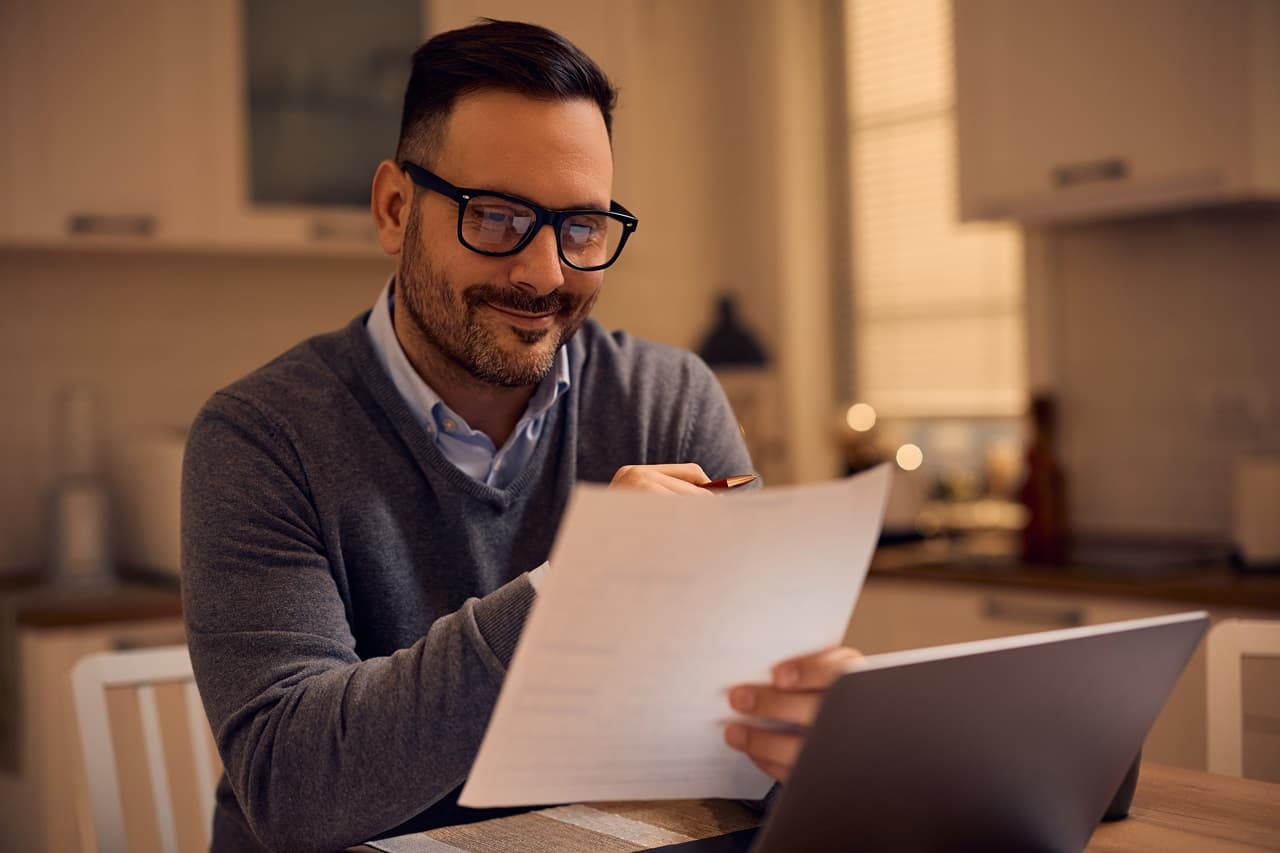 homem usando óculos de grau, sentado em frente a uma mesa com notebook, segurando uma folha de papel nas mãos.