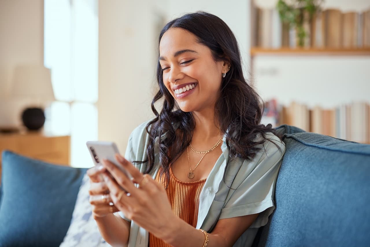 Mulher sorrindo, sentada em um sofá com celular na mão